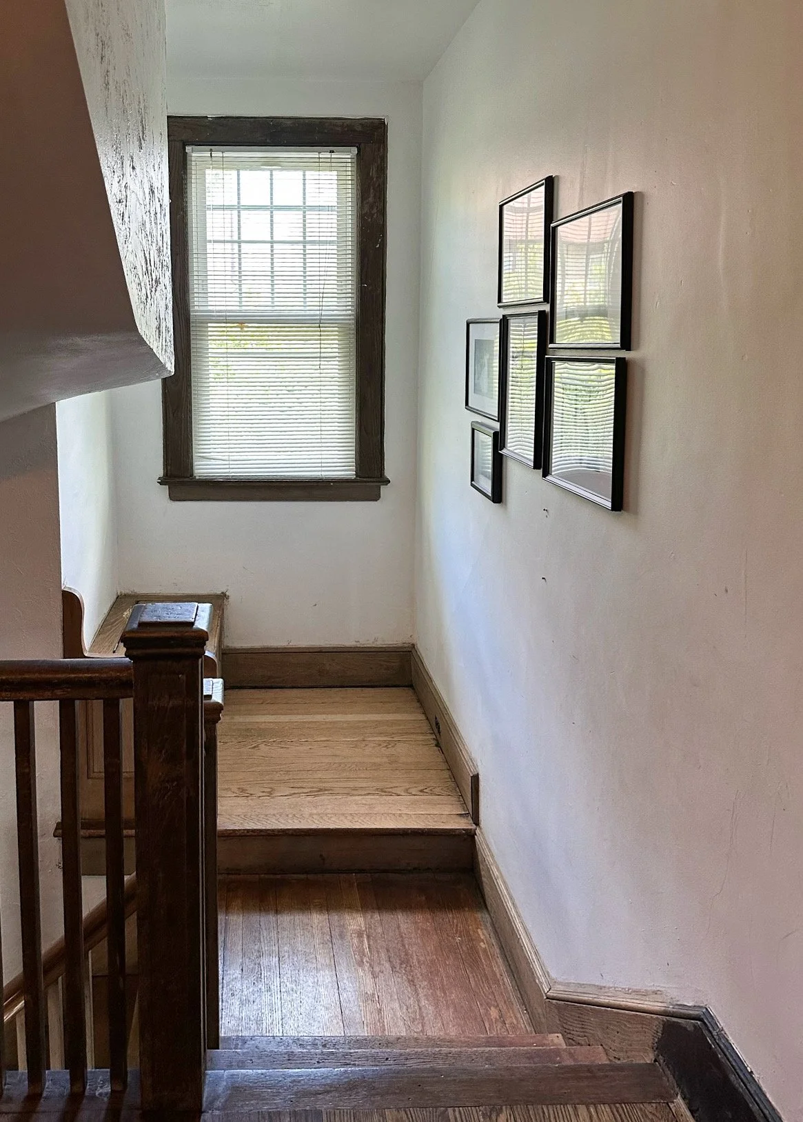 A small landing at the top of a staircase with a window, framed with dark wood, and a collection of five framed pictures on the wall.