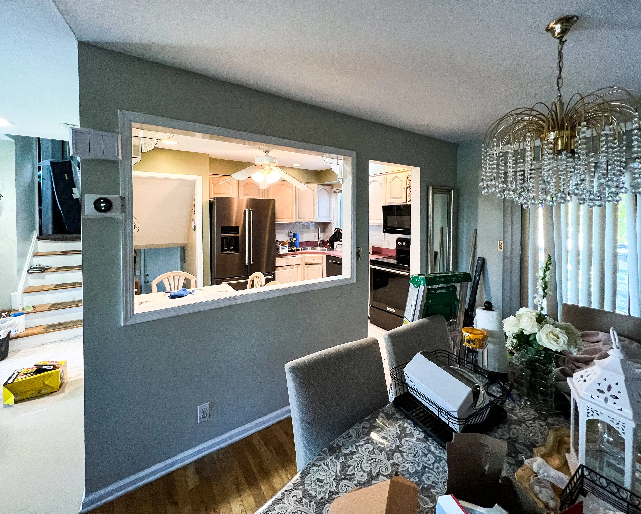 View of a dining area with a chandelier and a living room, with a view into the kitchen through a large open window. The kitchen contains a black refrigerator, wooden cabinets, and a white ceiling fan. The dining table is cluttered with various items
