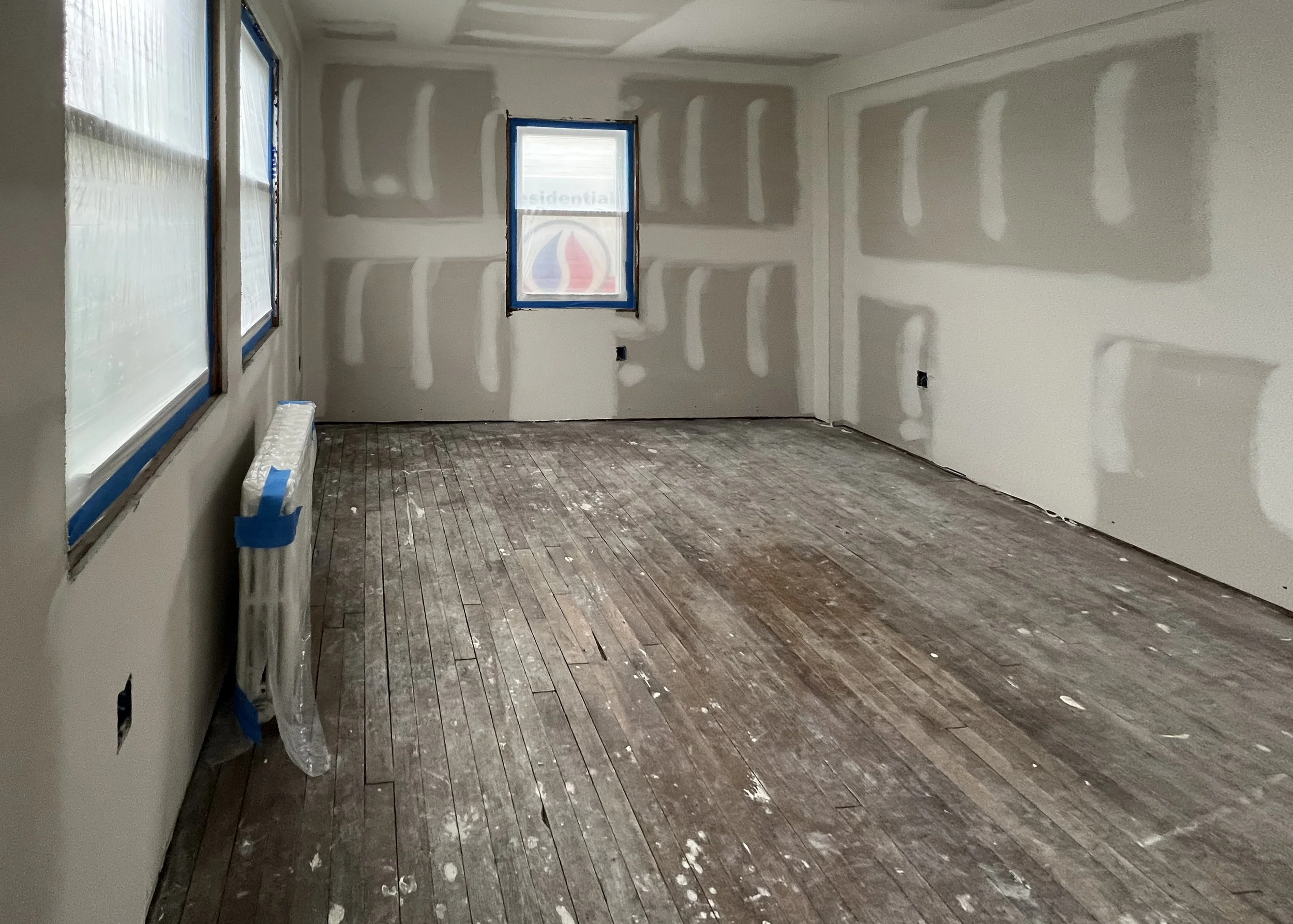 Empty room under construction with drywall installed and taped, three windows covered with plastic, and a wooden floor with paint splatters.