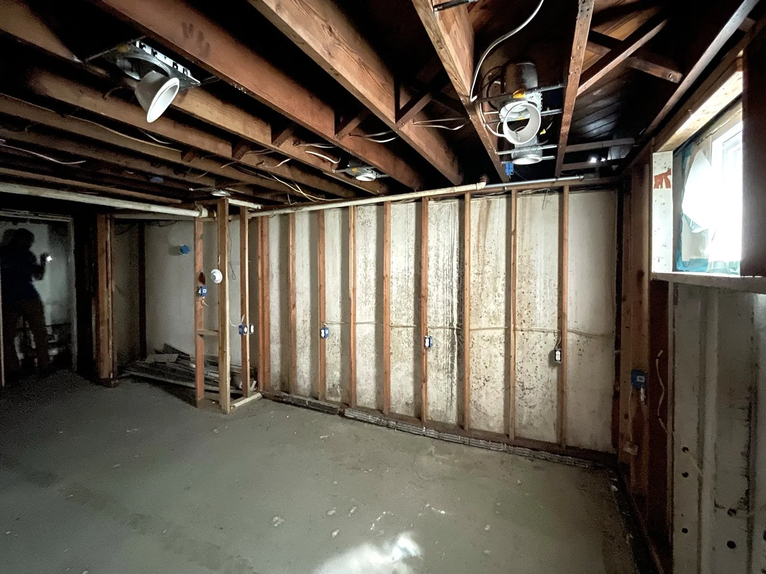 Empty room under construction with exposed wooden framing, electrical outlets, and a small window, with a person taking a photo in the background.