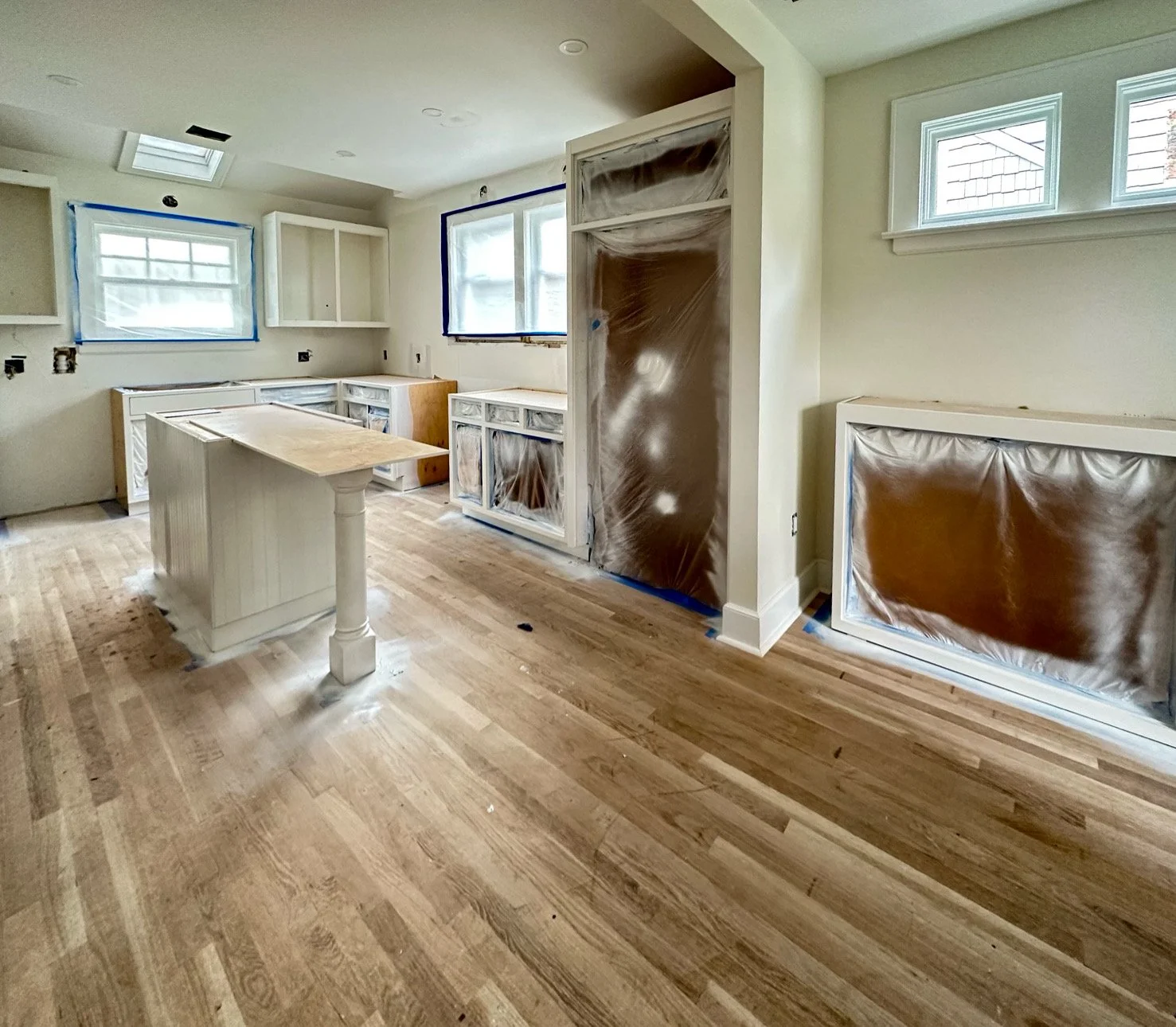 Kitchen under renovation with wooden floors, white cabinets, window coverings, and plastic coverings on cabinets and door.