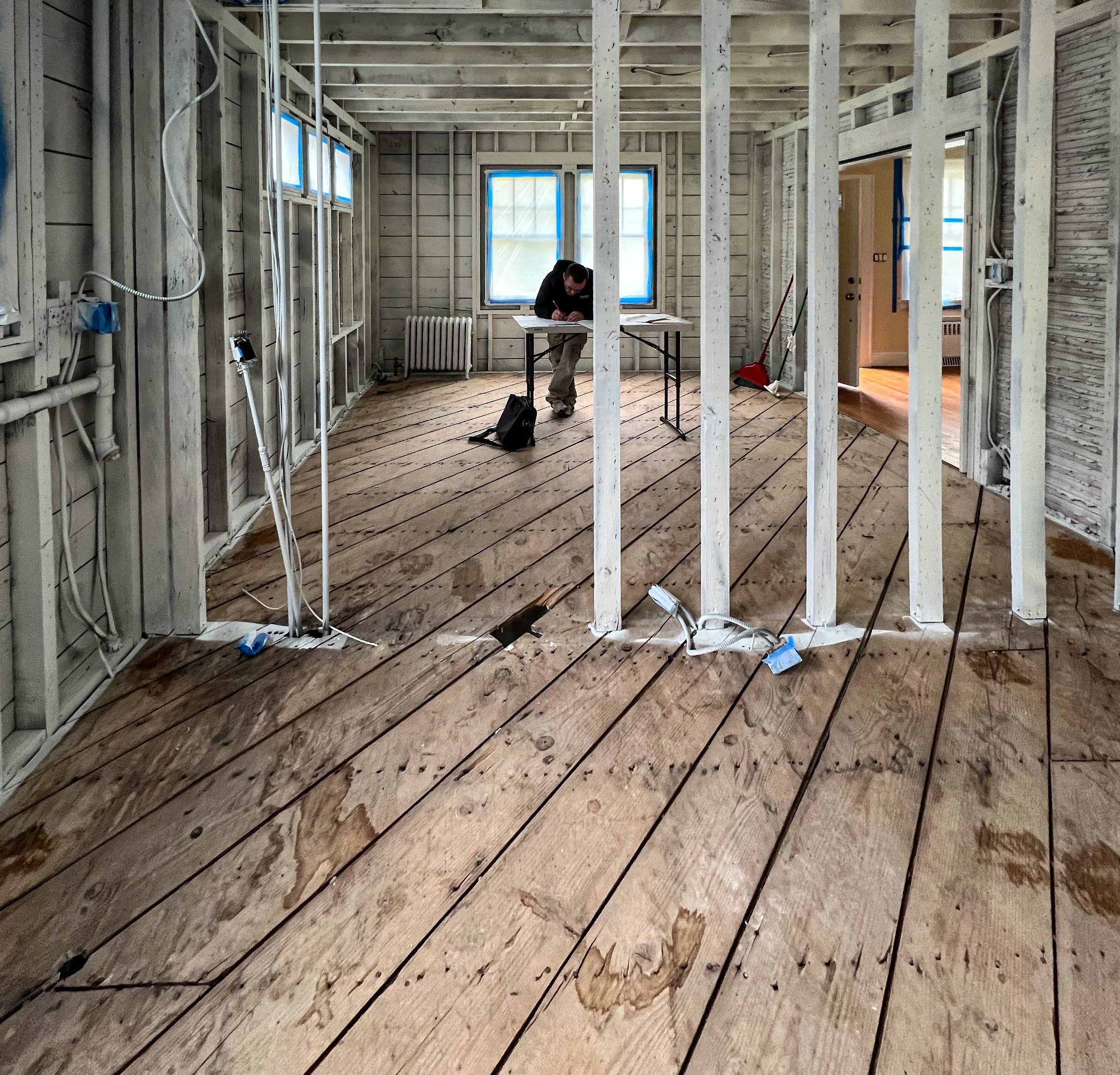 Interior of a house under renovation with exposed framing, electrical wiring, and hardwood floors; a person charts or designs at a table near windows.