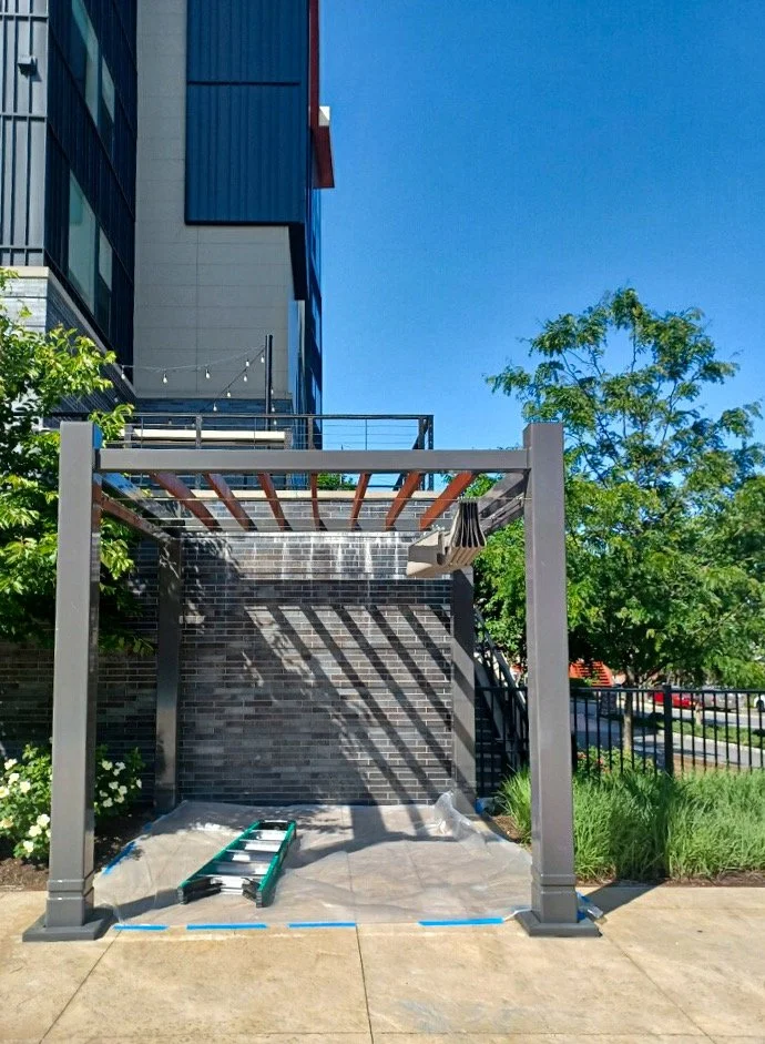 A partially constructed pergola with metal posts and wooden beams in an outdoor area, with a brick wall, trees, and a building in the background under a clear blue sky.