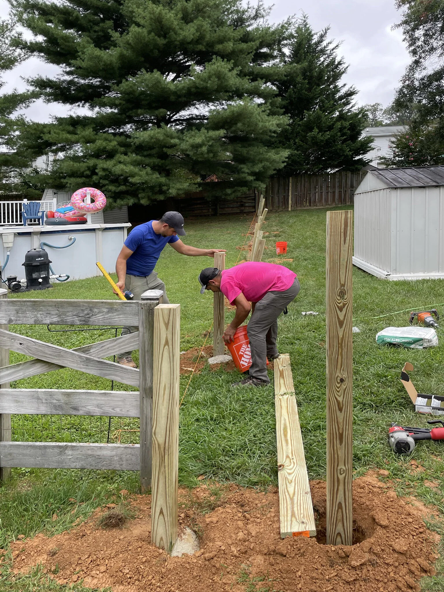 Two men building a wooden fence in a backyard with trees, a shed, and a pool with toys.