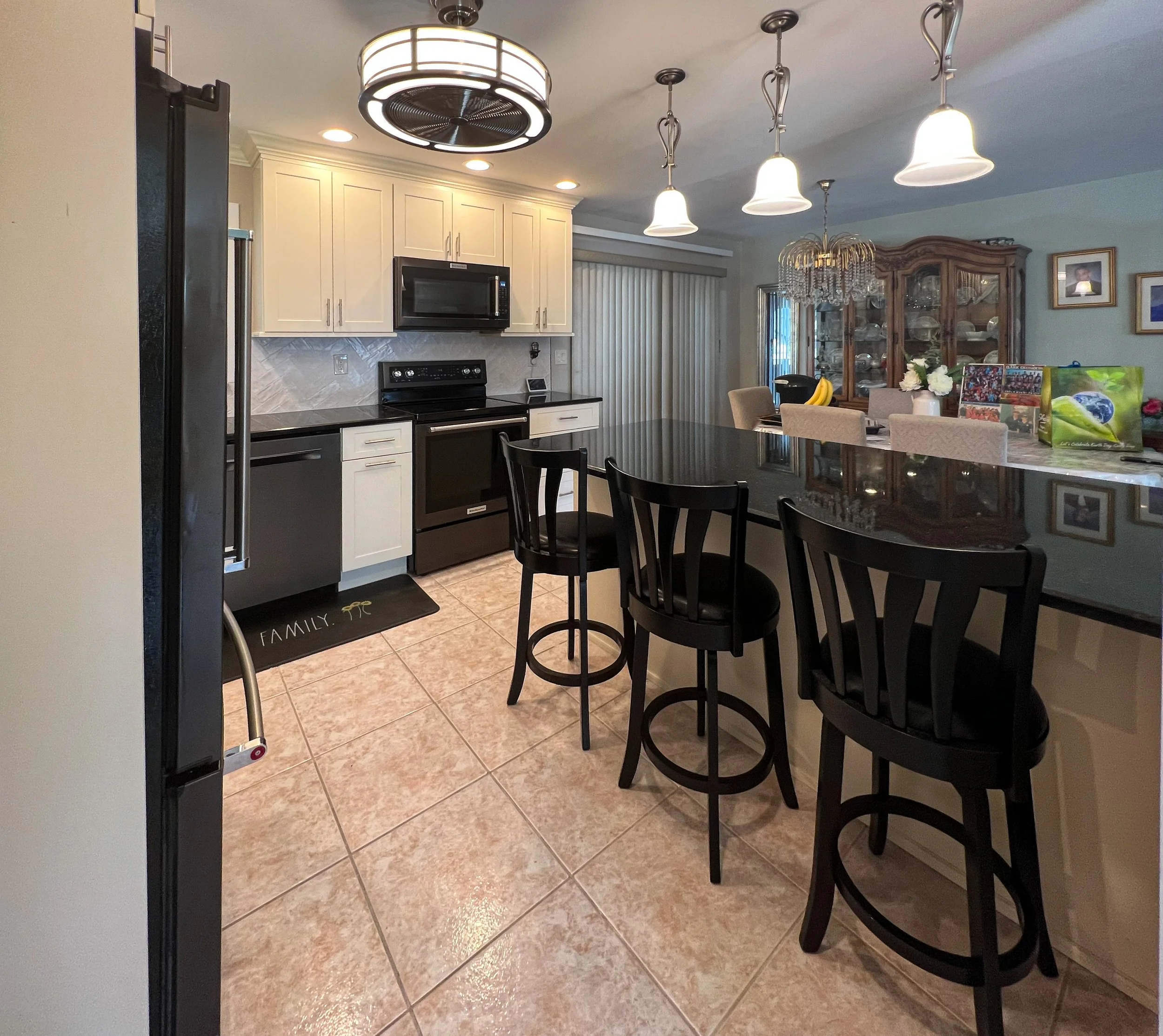Kitchen with white cabinets, black appliances, tiled floor, and a dark countertop bar with black chairs. There are pendant lights and a ceiling fan, and a dining area with a glass-front cabinet and chairs in the background.