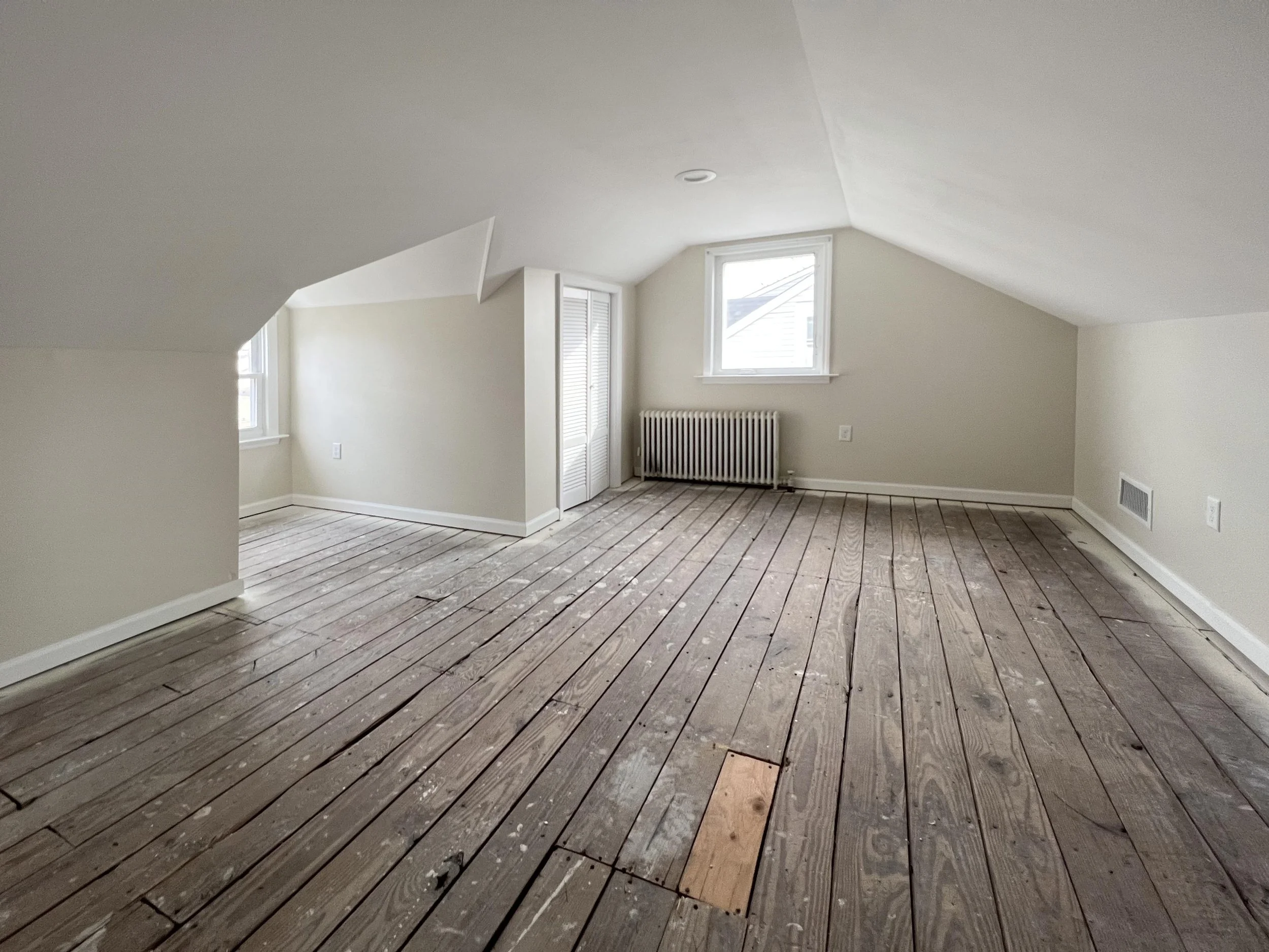 Empty attic room with unfinished wooden flooring, white walls, two windows, a radiator, and built-in closet.