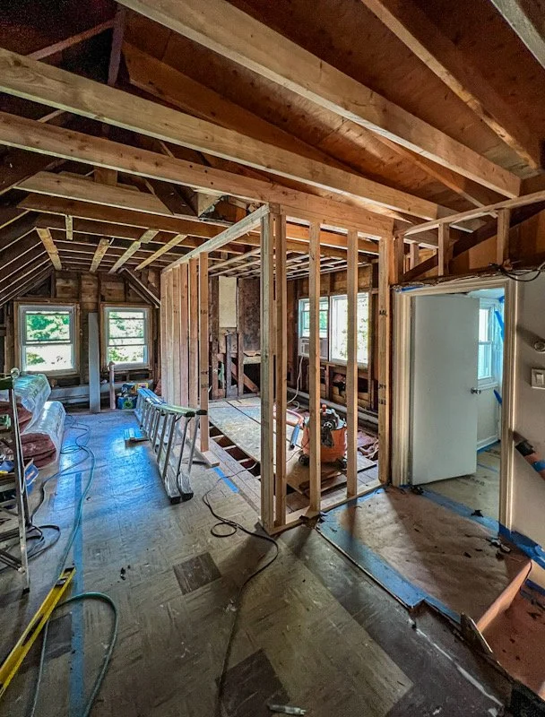 Interior view of a house under construction, showing exposed wooden framing, construction tools, and partially finished walls and floor.