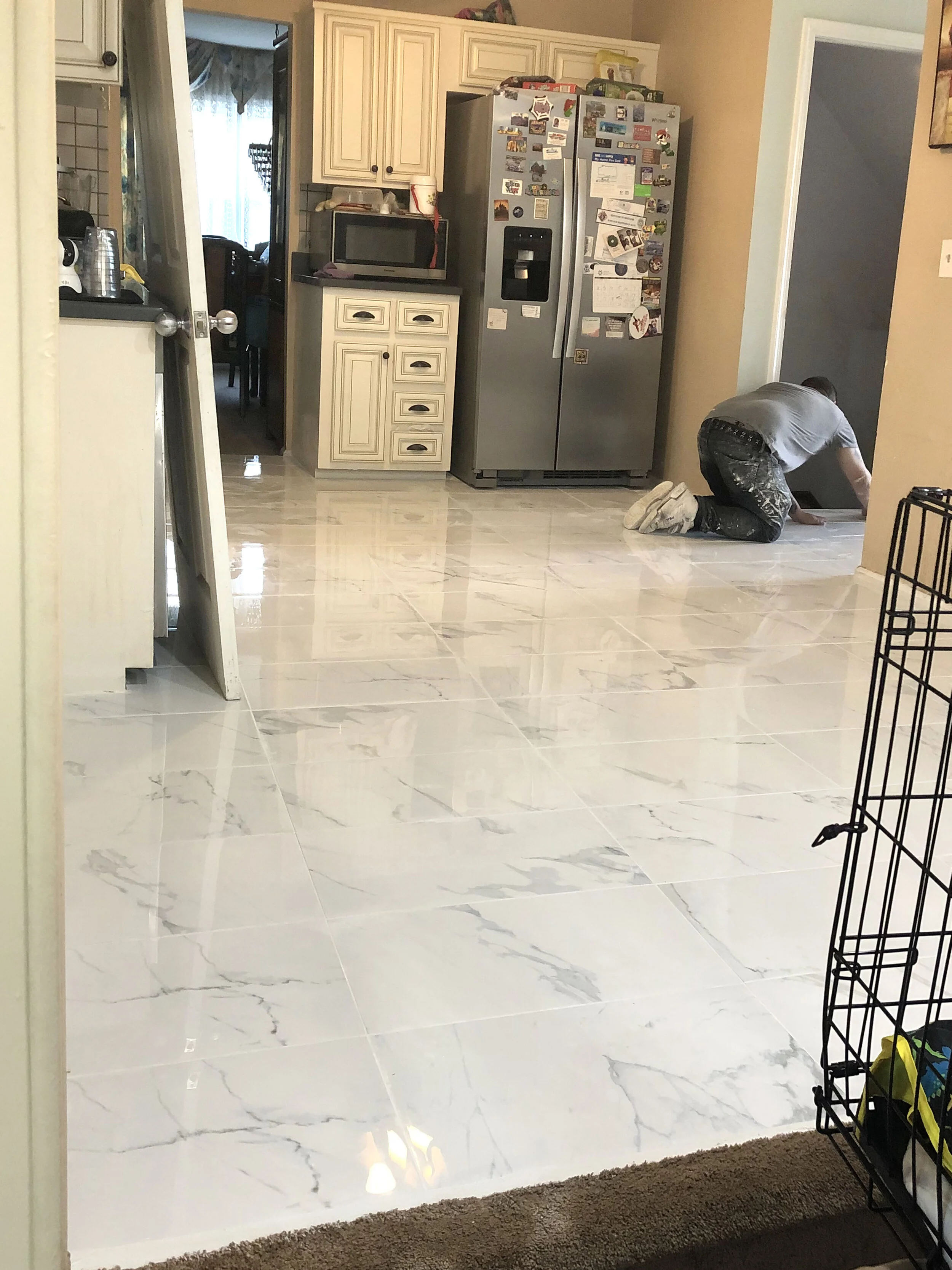 A person kneeling on kitchen floor installing or cleaning tiles, with kitchen appliances and cabinets in the background.