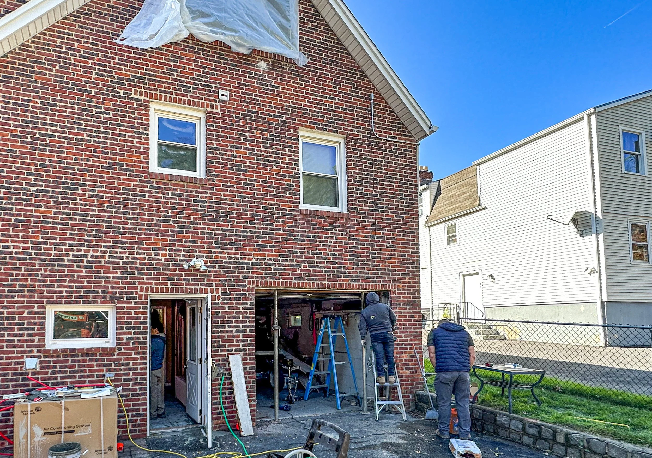 Construction workers working on the back of a two-story brick house with a garage, ladders, and tools, while a neighboring house is visible to the right under a clear blue sky.