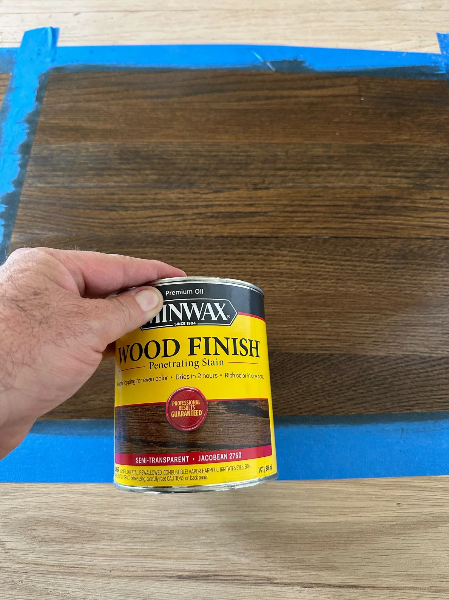 A hand holding a can of Minwax Wood Finish penetrating stain in the color Jacobean 2750, with a piece of wood that has been stained with the same product placed on a surface covered with blue painter's tape.