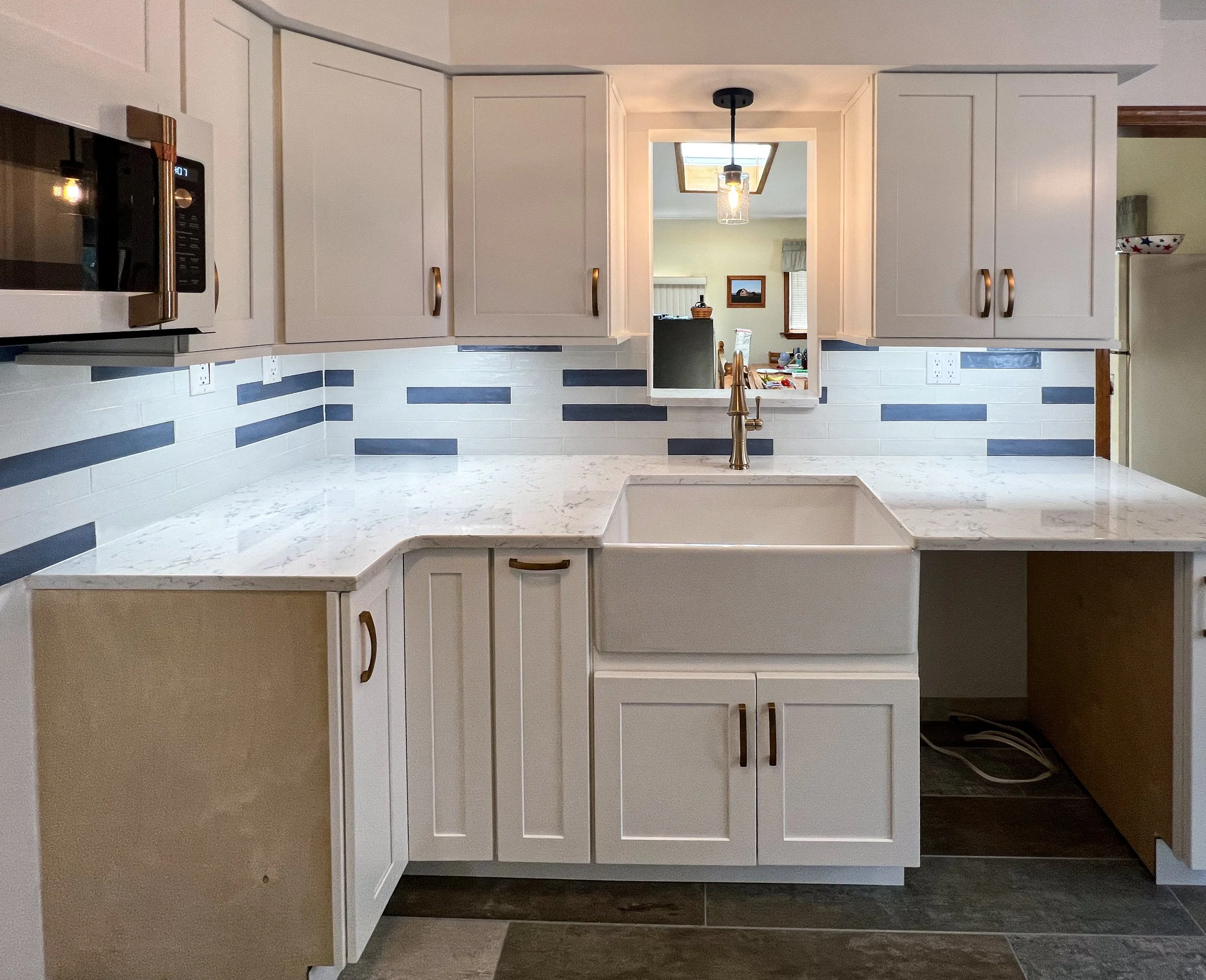 Kitchen with white cabinets, marble countertop, a white farmhouse sink, and a backsplash with horizontal gray and white tiles. There is a small window opening to a dining area visible in the background.