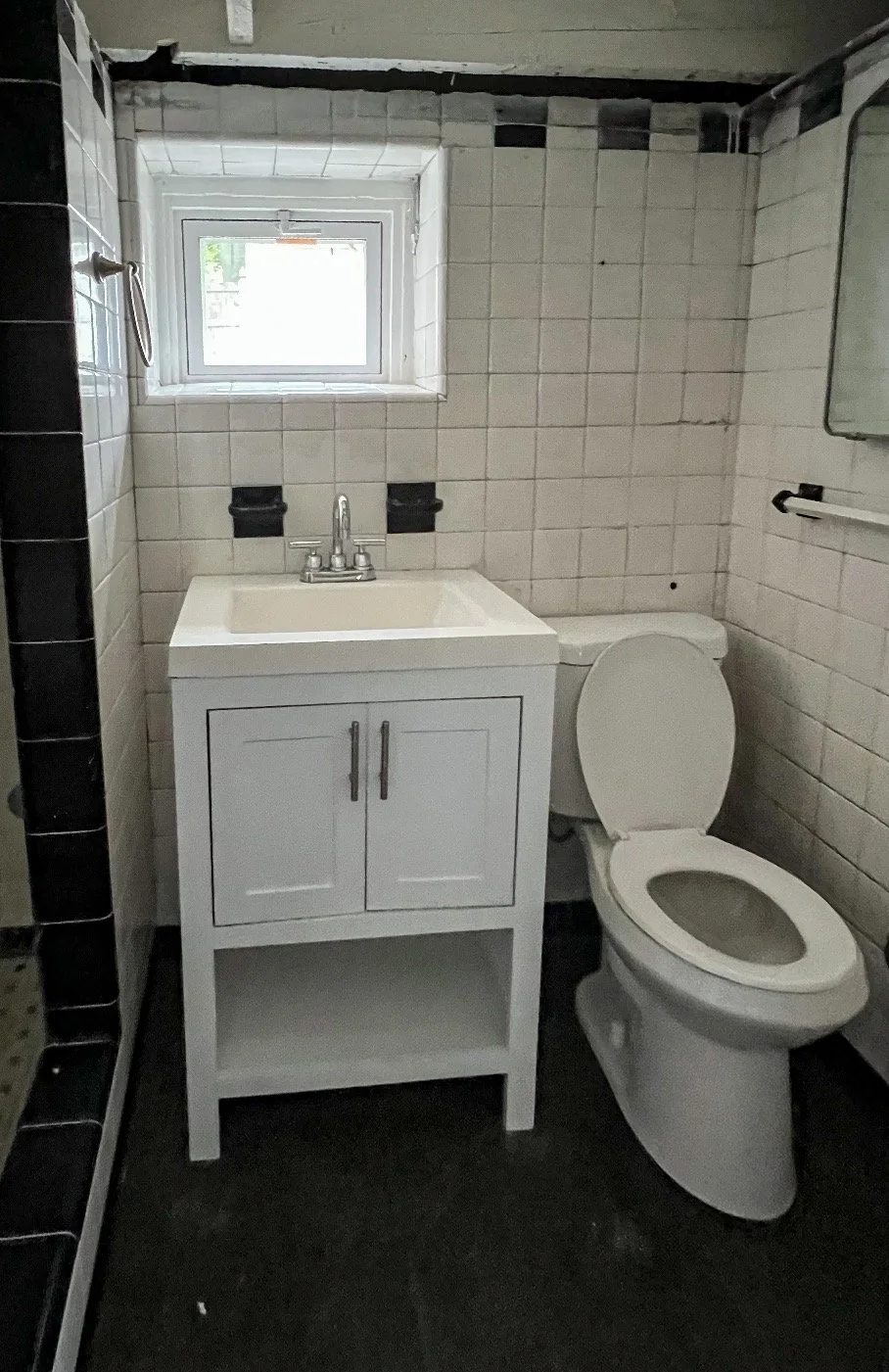 Small bathroom with a white sink cabinet, toilet, small window, tiled walls, and part of a shower area.