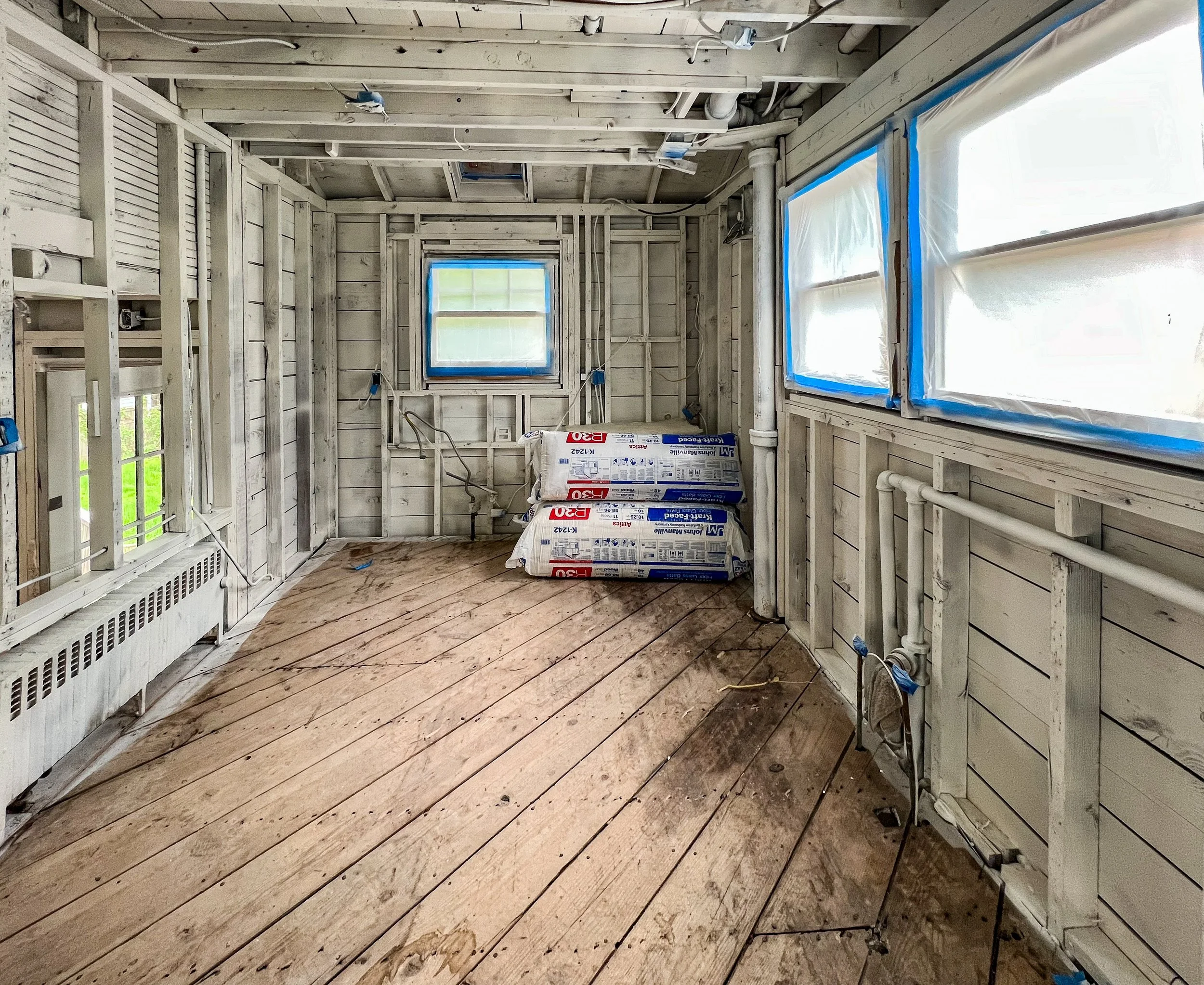 Room under construction with exposed wooden framing, plastic-wrapped windows, and stacks of insulation bags on the floor.
