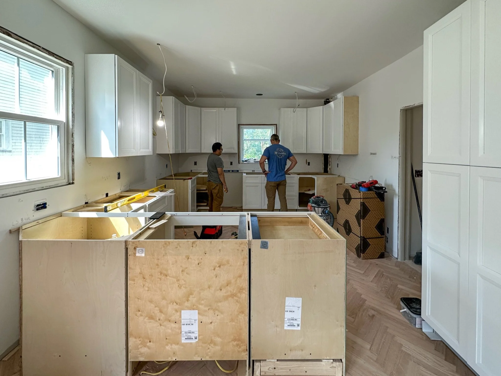 Kitchen under construction with two workers, white cabinets, and construction materials.