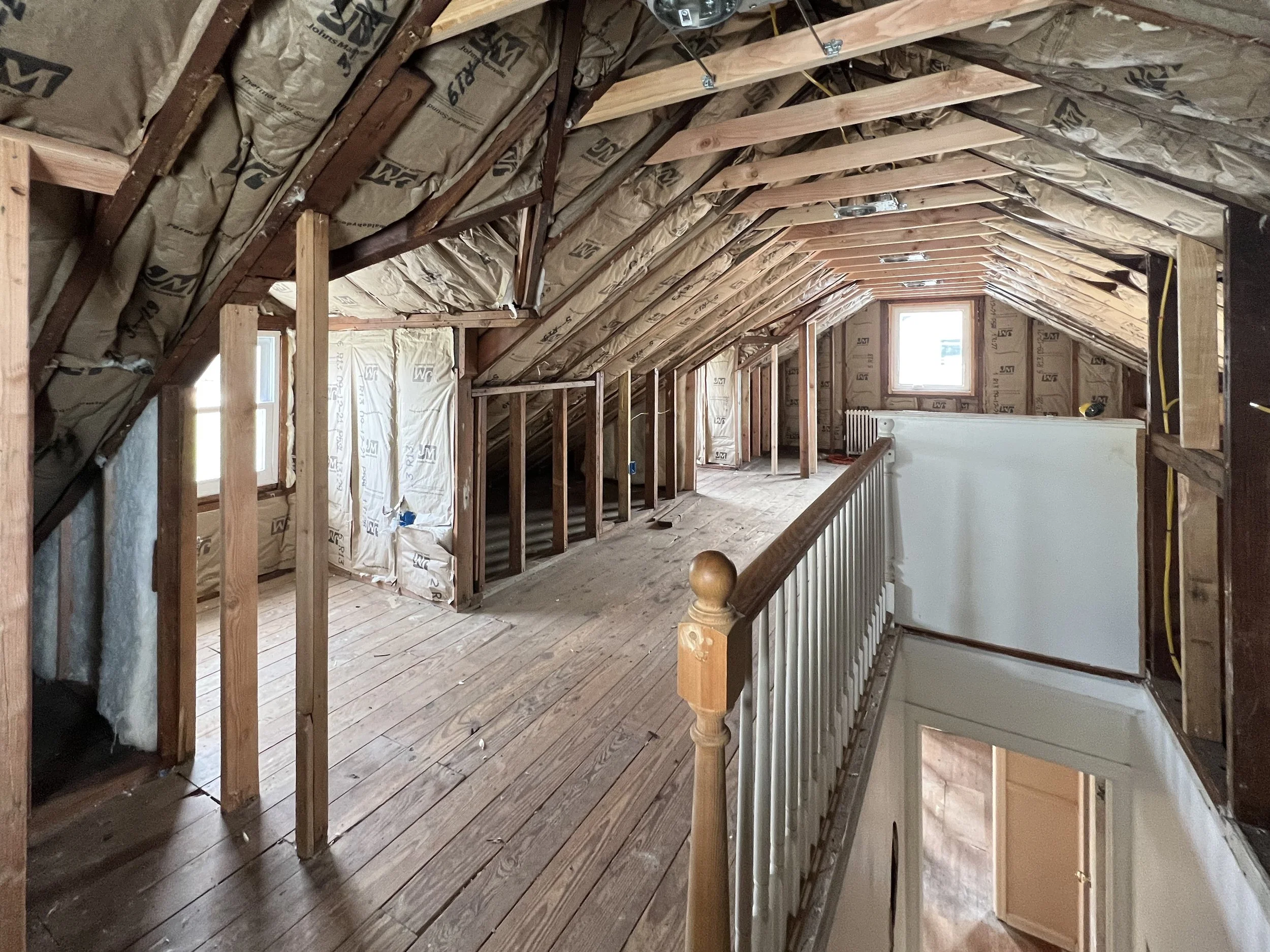 Attic renovation in progress with exposed wood framing, insulation, and a wooden floor. Small windows at the end and natural light visible.