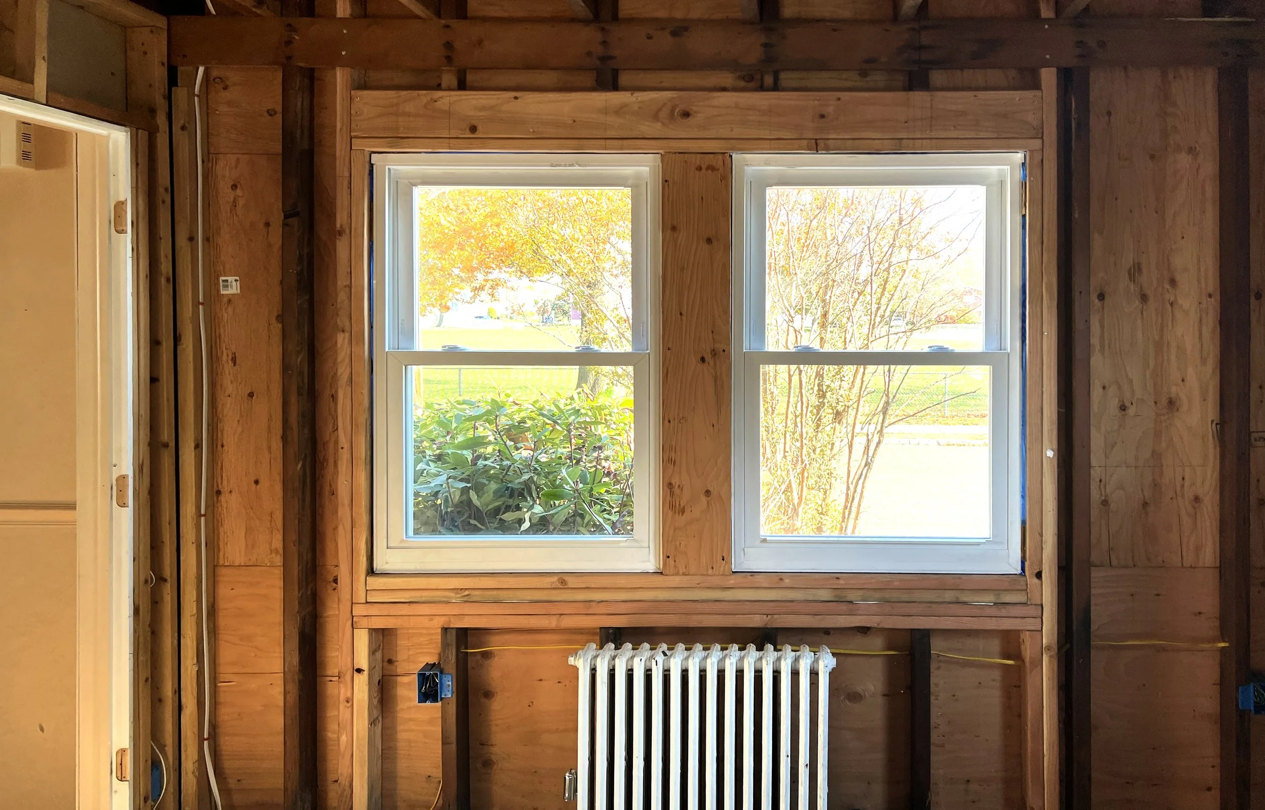 Interior view of a partially finished room with two double-hung windows, wooden framing, and a white radiator.