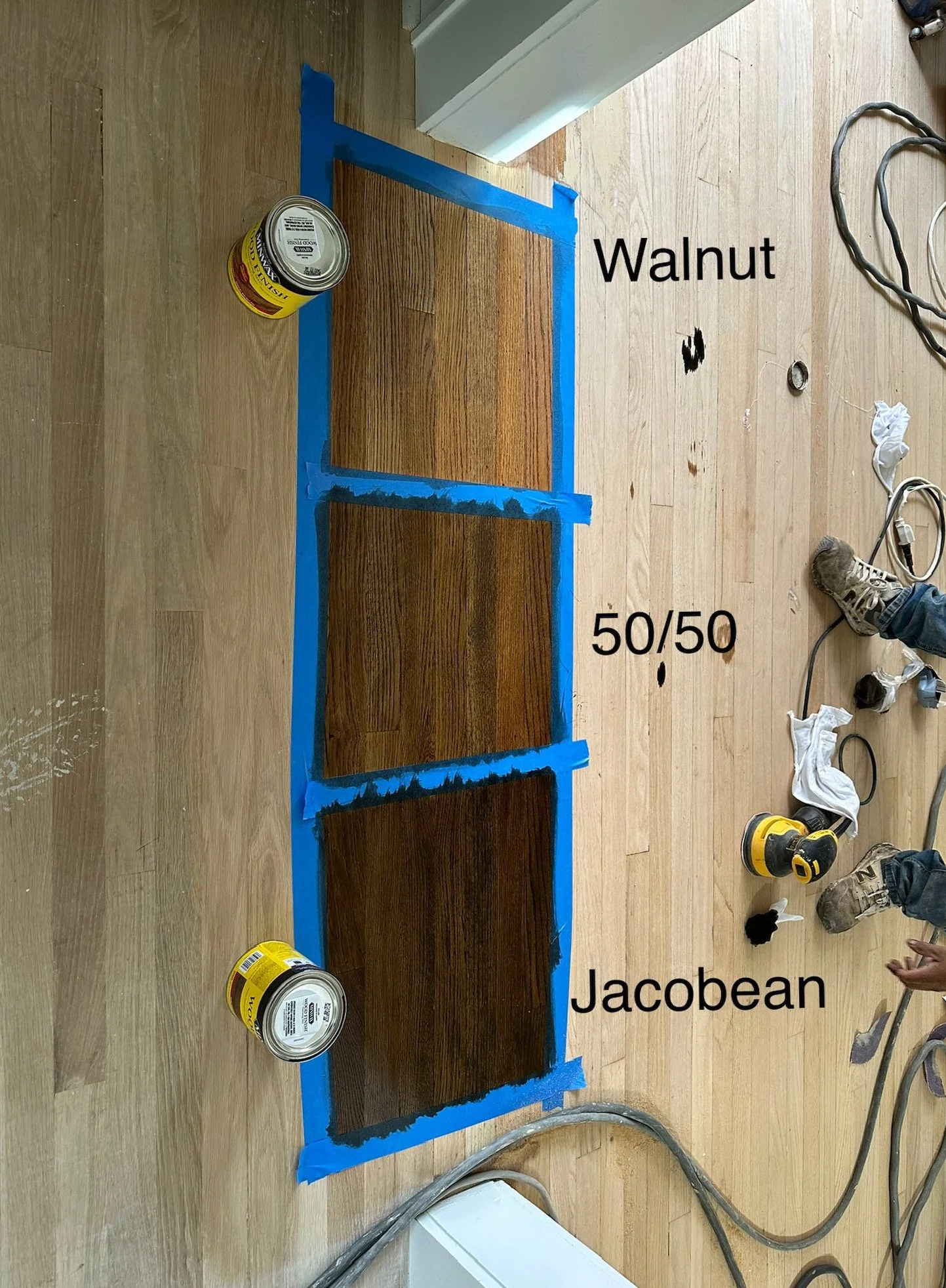 Three wooden floor samples labeled Walnut, 50/50, and Jacobean, taped on a floor with painter's tape. There are cans of stain or finish, some tools, and shoes around the samples. The labels indicate different wood stain colors.