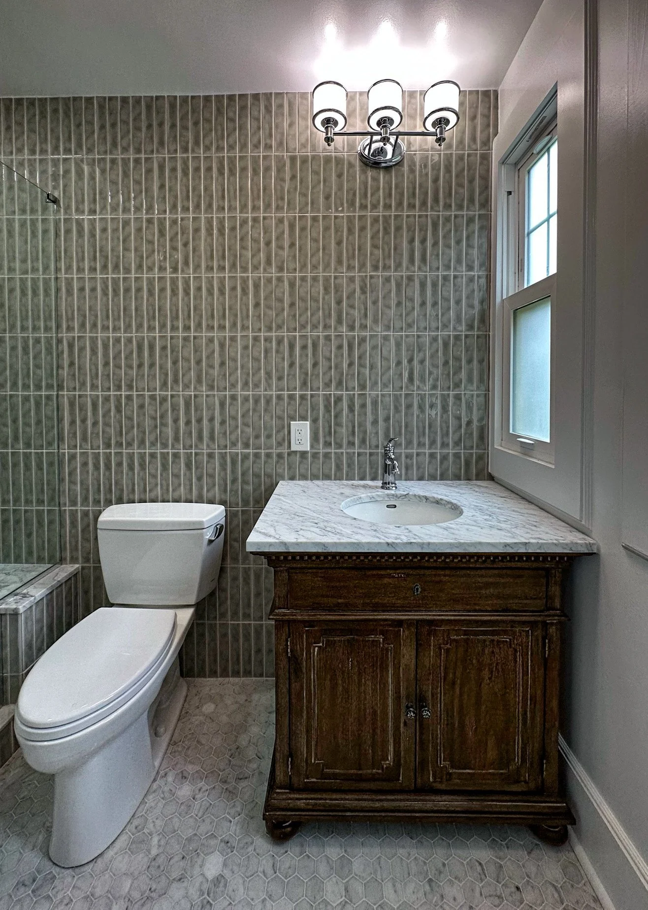 Bathroom with a toilet, wooden vanity with marble top, small sink, wall-mounted light fixture, window, and tiled wall and floor.
