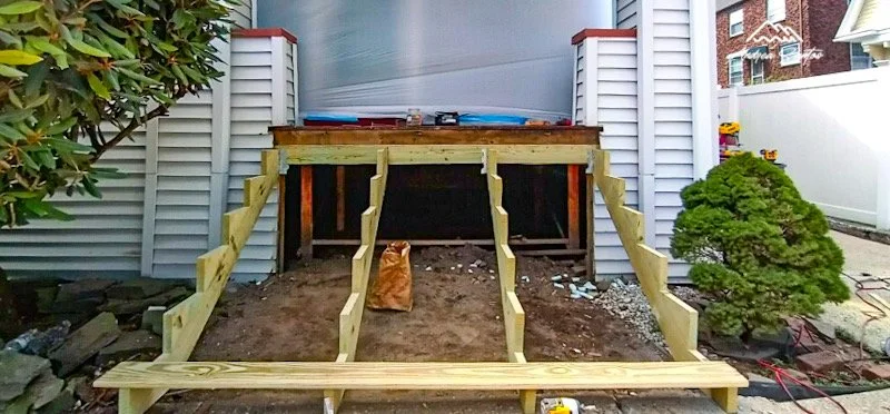 Partially built wooden staircase leading to a raised platform at the back of a house, with gardening tools and materials nearby, and a bush on the right side.