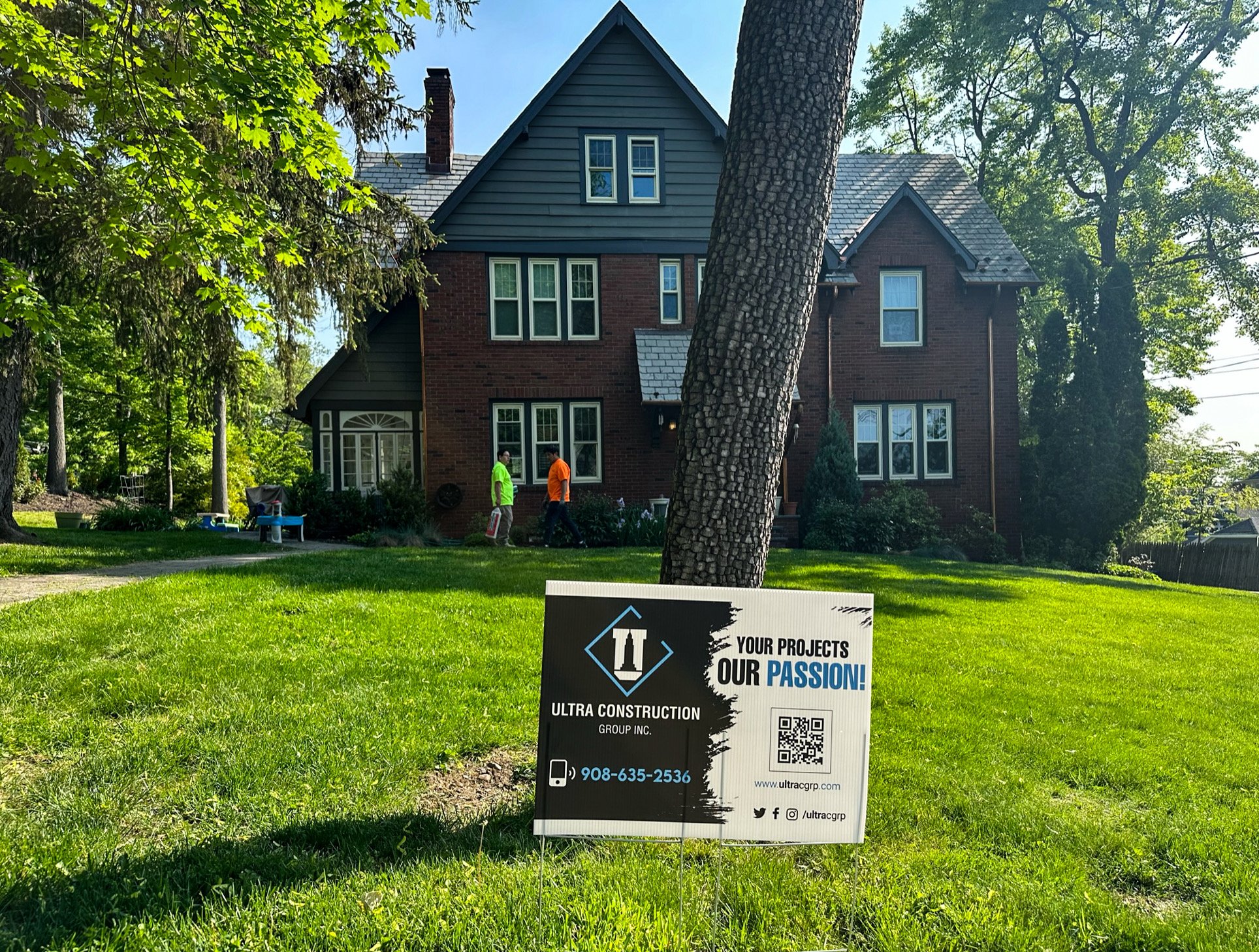 A large two-story brick house with a gray roof, surrounded by trees and a well-maintained lawn. Two workers are walking on the lawn, and a sign in the foreground shows the logo, contact information, and slogan for Ultra Construction Group Inc.