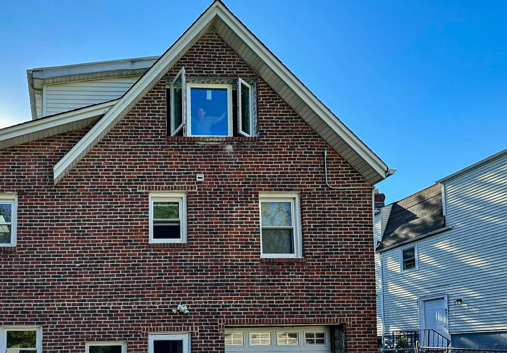 A red brick house with a gable roof and white trim. There is an open window on the upper level, and two smaller windows on the lower level, with a garage door at the bottom. The sky is clear and blue.