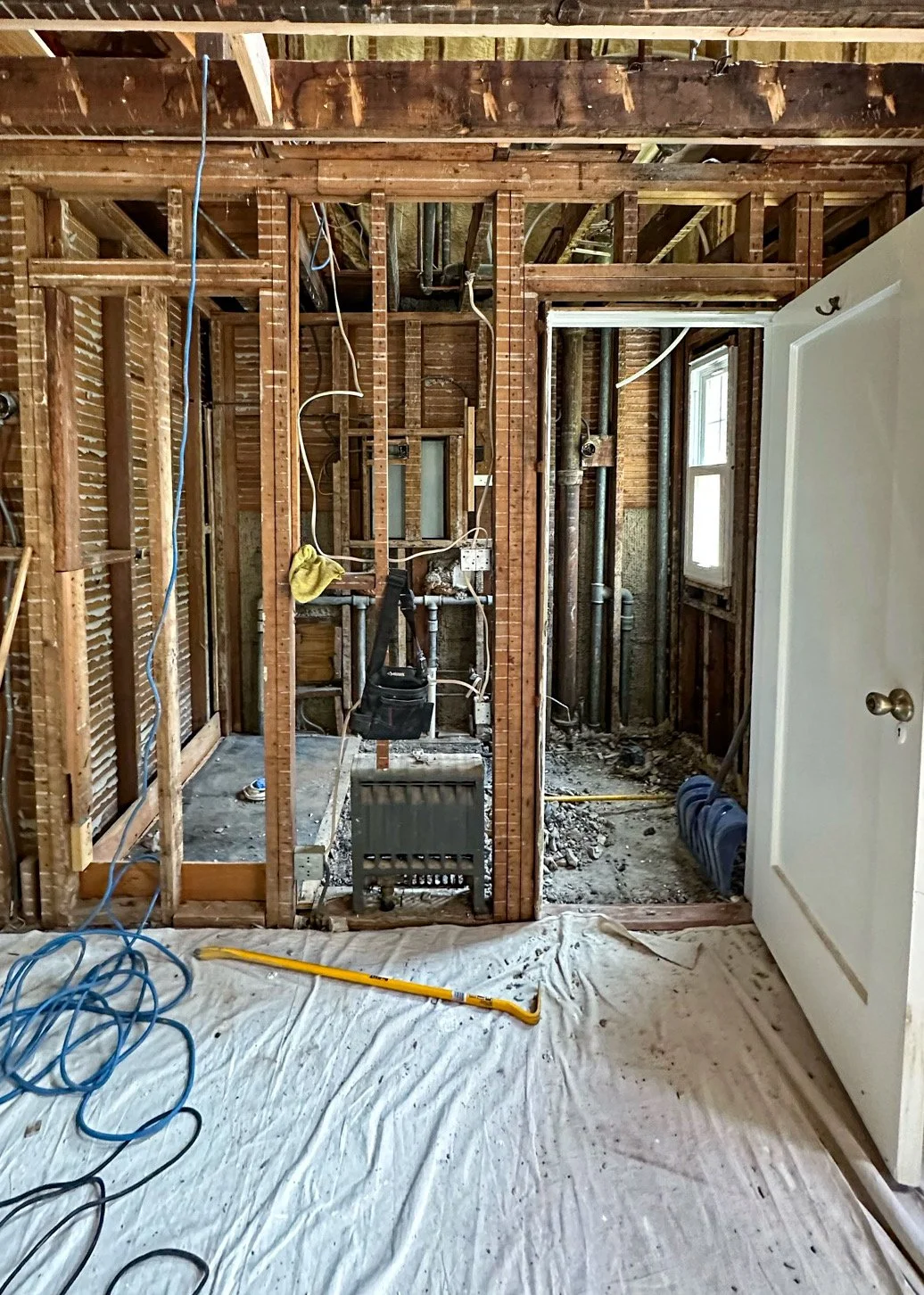 Interior view of a room under renovation, showing exposed wooden framing, electrical wiring, and plumbing pipes. There is a window on the right, construction tools on the floor, and a partially removed wall.