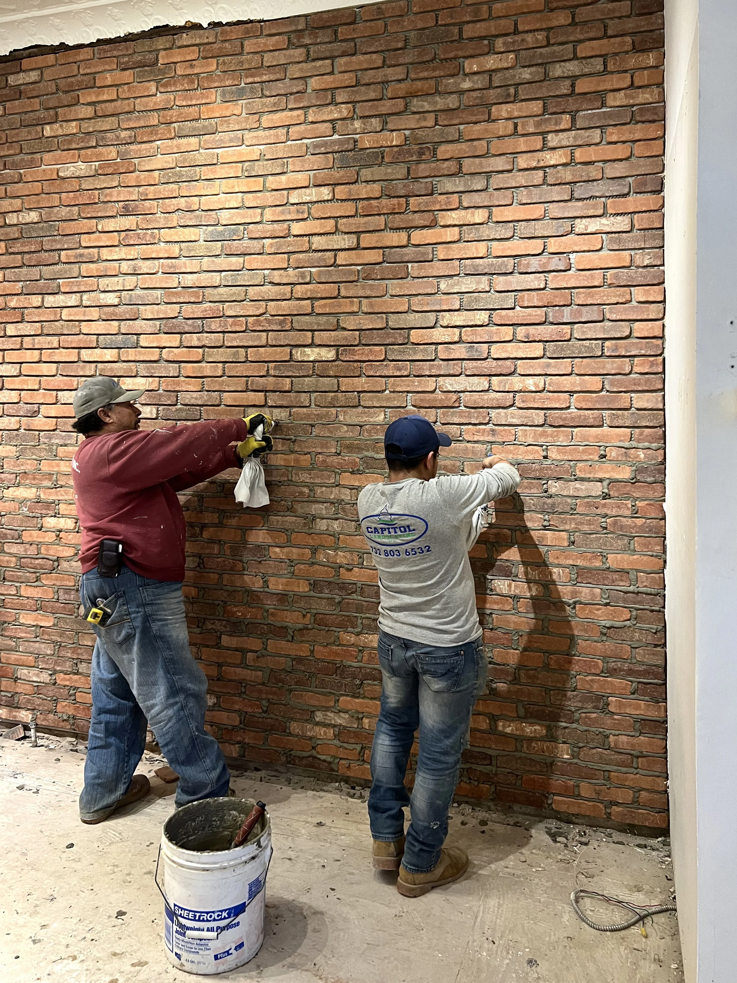 Two workers installing bricks on a wall, one holding a brick and the other applying mortar. There's a bucket of mortar and construction tools nearby.