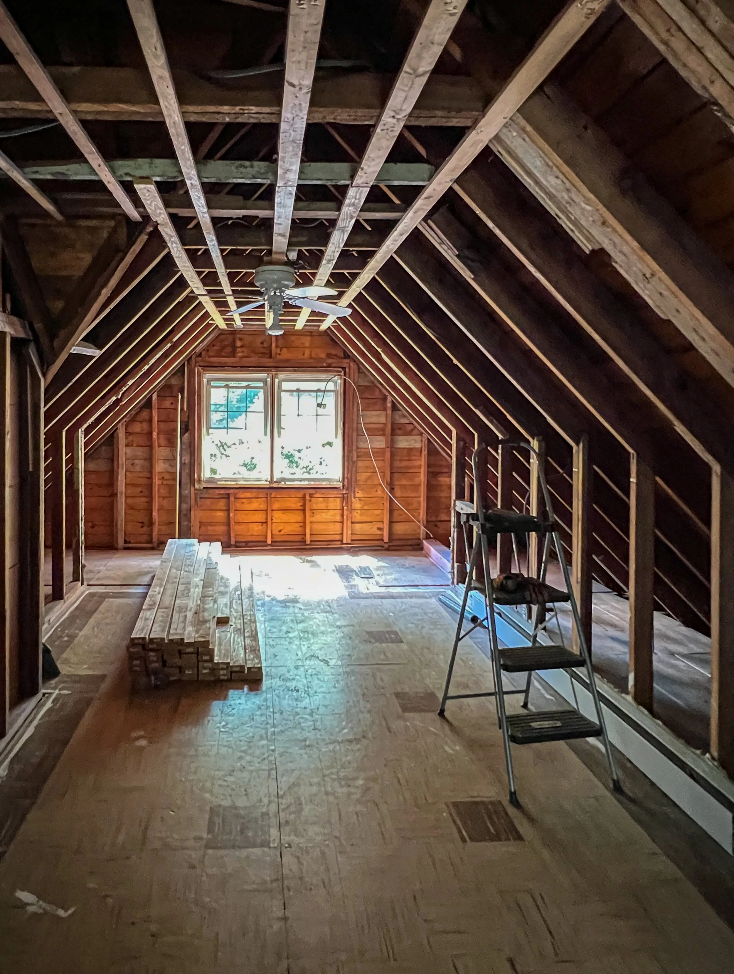 Attic room under renovation with exposed wooden framing, a window at the far end, a ceiling fan, a stepladder, piles of wooden planks, and unfinished flooring.