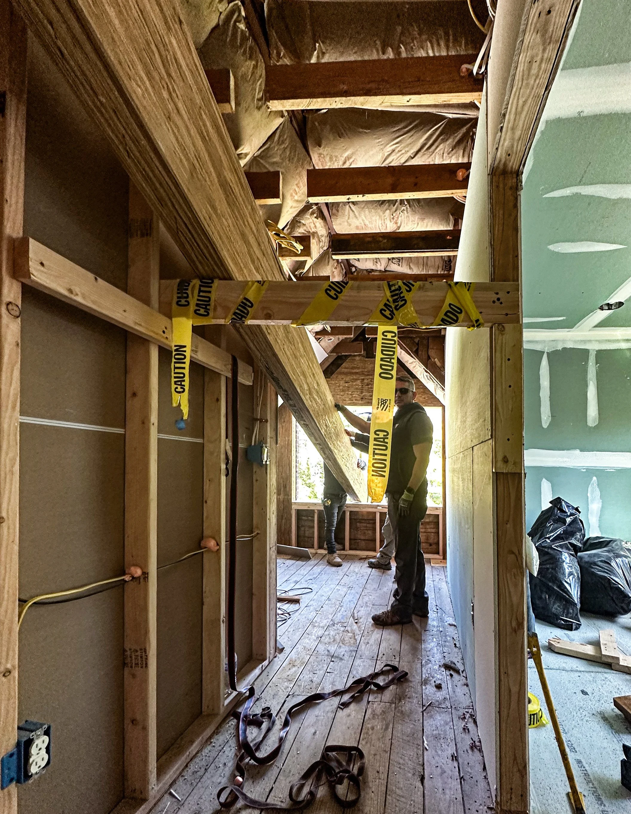 Construction workers in a partially built room with wooden framing, insulation, and caution tape.