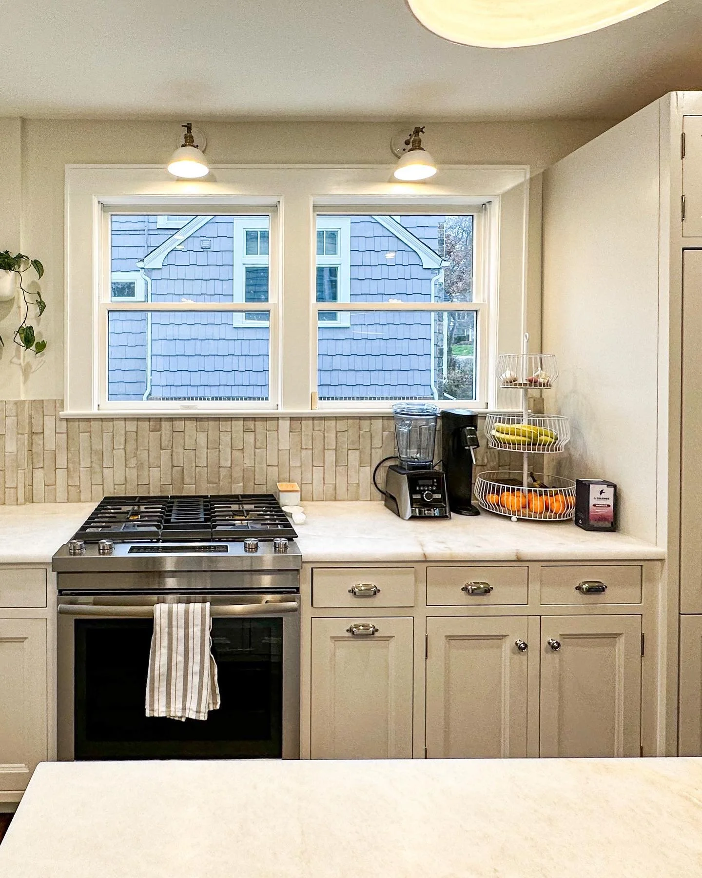 A kitchen with a gas stove, white cabinets, a beige countertop, a fruit basket, blender, and a window with a view of a blue house exterior.