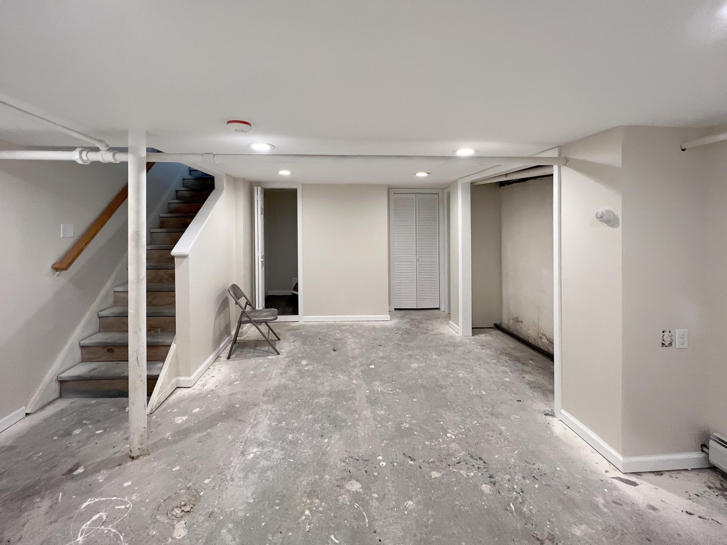 Basement with unfinished concrete floor, white walls, a staircase with wooden steps and rail, a metal folding chair, and white closet doors, with visible ductwork on the ceiling.