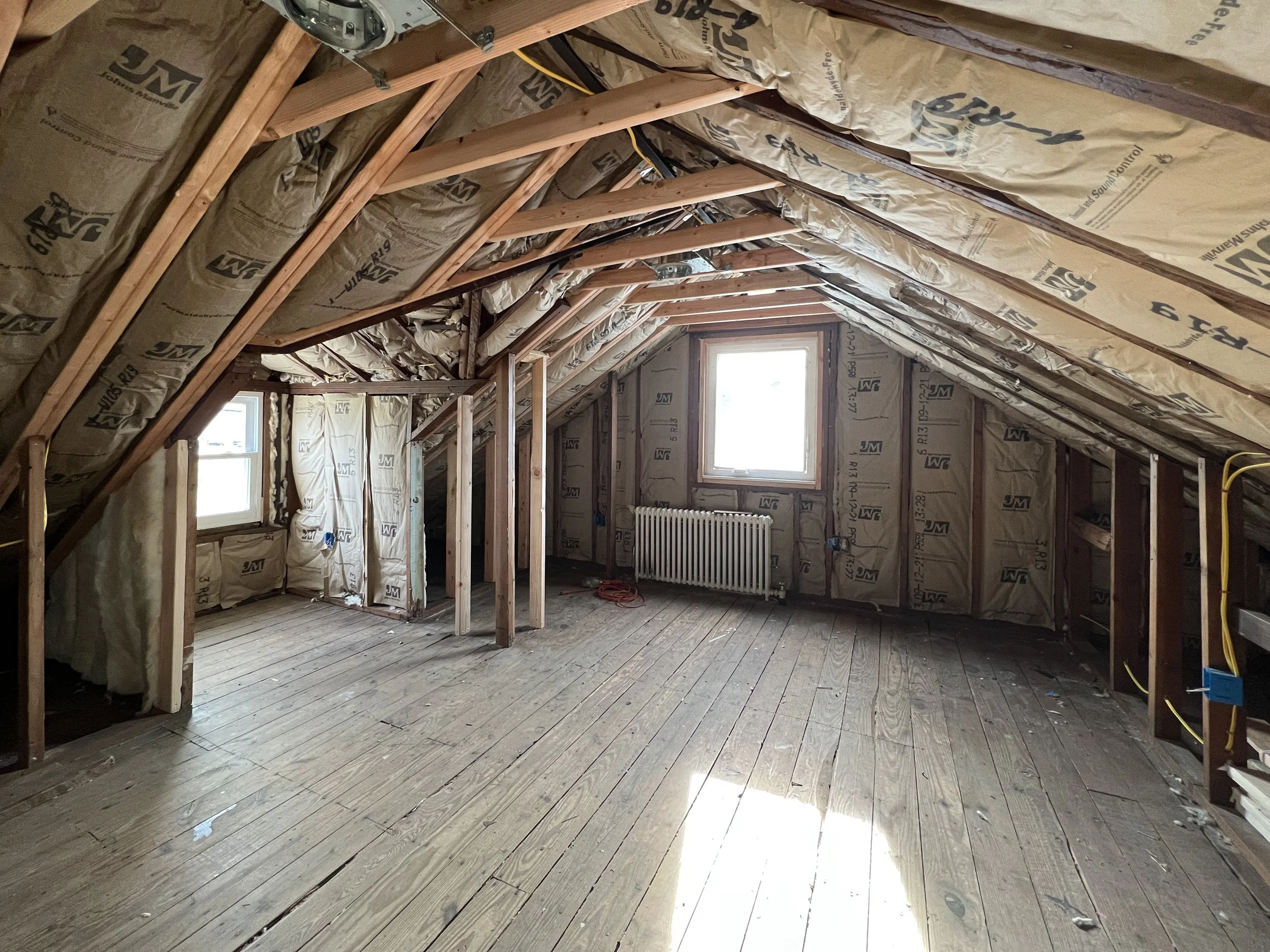 An unfinished attic with insulation, wooden beams, and two windows, with a radiator beneath one window.