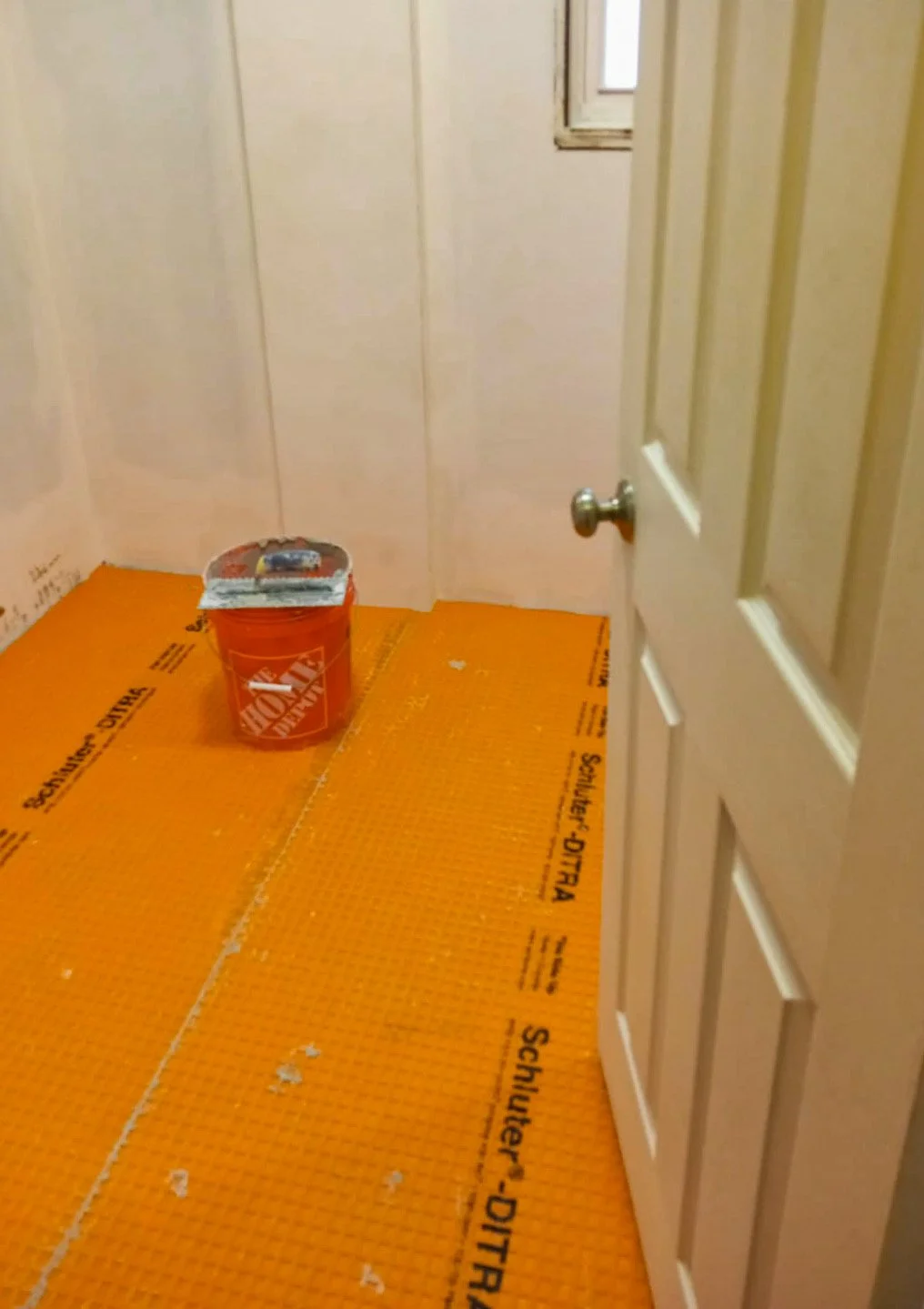 A small, partially finished room with light-colored walls, an open door, and a window. The floor is covered with orange SCHLUTER-DITRA underlayment, and there is a red Home Depot bucket on the floor near the window.