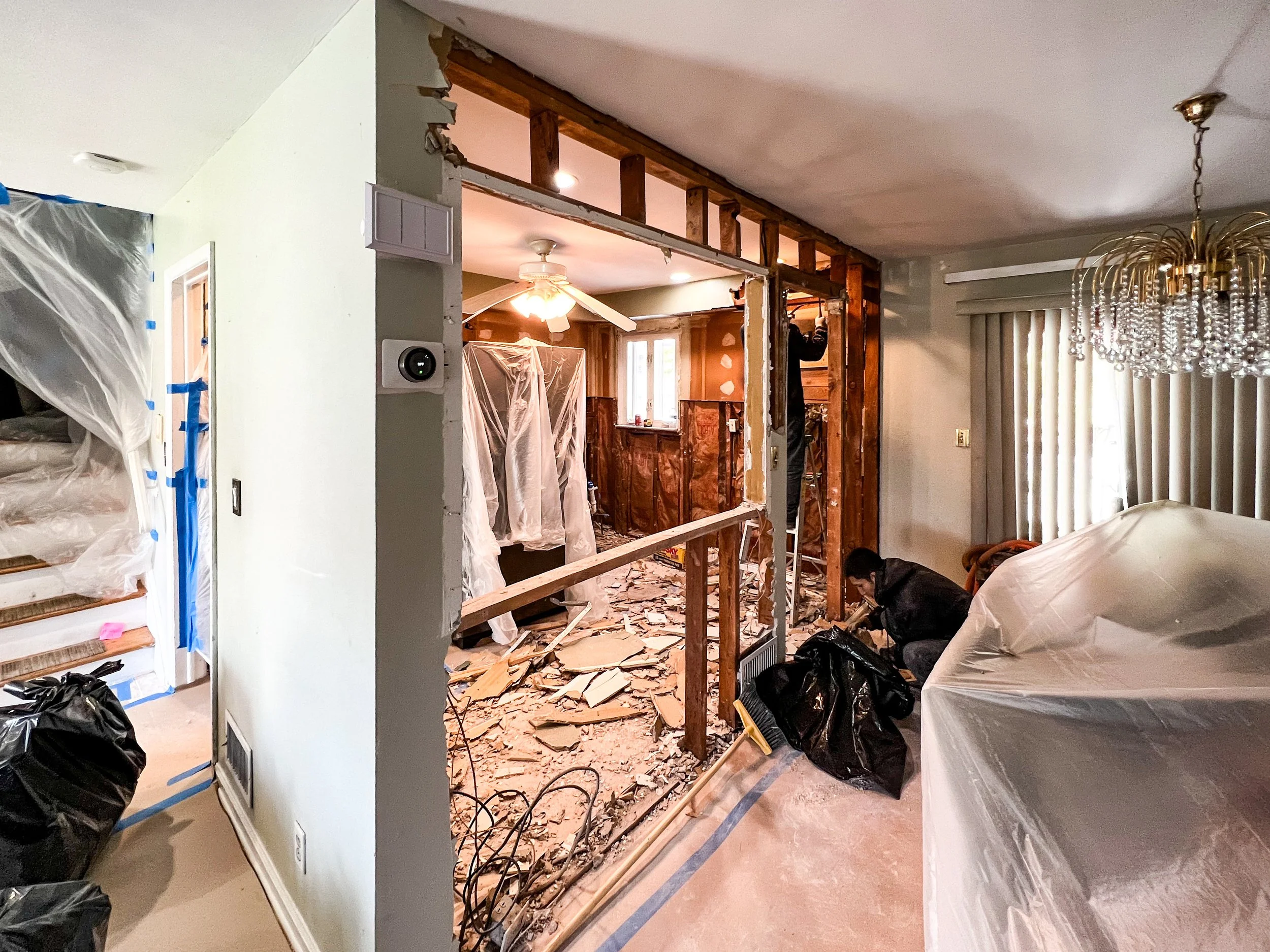 Room undergoing renovation with exposed wooden framing, debris on the floor, and two workers dismantling a wall, with covered furniture and construction materials present.