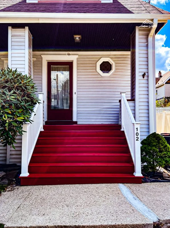 Front porch of a house with bright red stairs, white railings, a burgundy front door, an octagonal window, white siding, and a bush to the right.