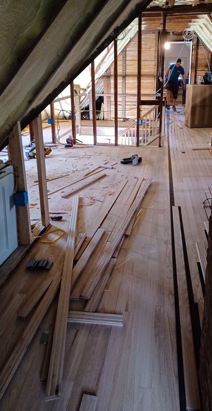 Interior of a house under renovation with wooden flooring, construction tools, and a man working in the background.