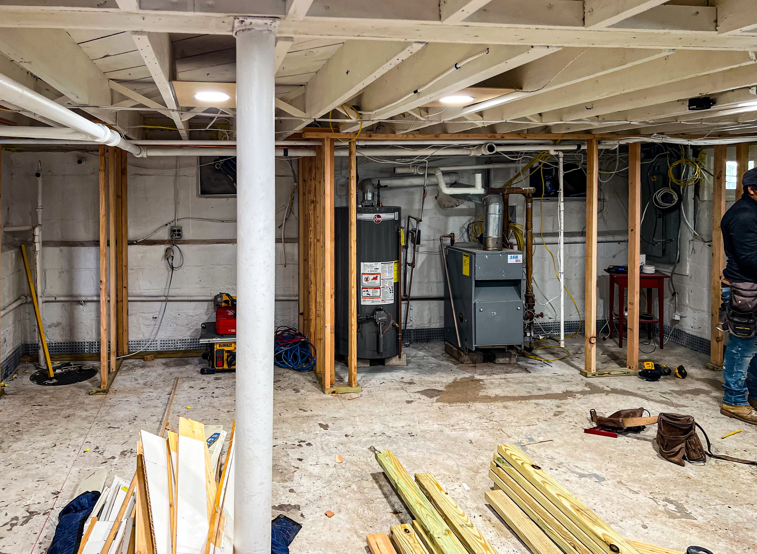 Construction site of a basement with exposed pipes and wooden framing, tools and materials scattered on the floor, and a partially installed furnace and water heater.