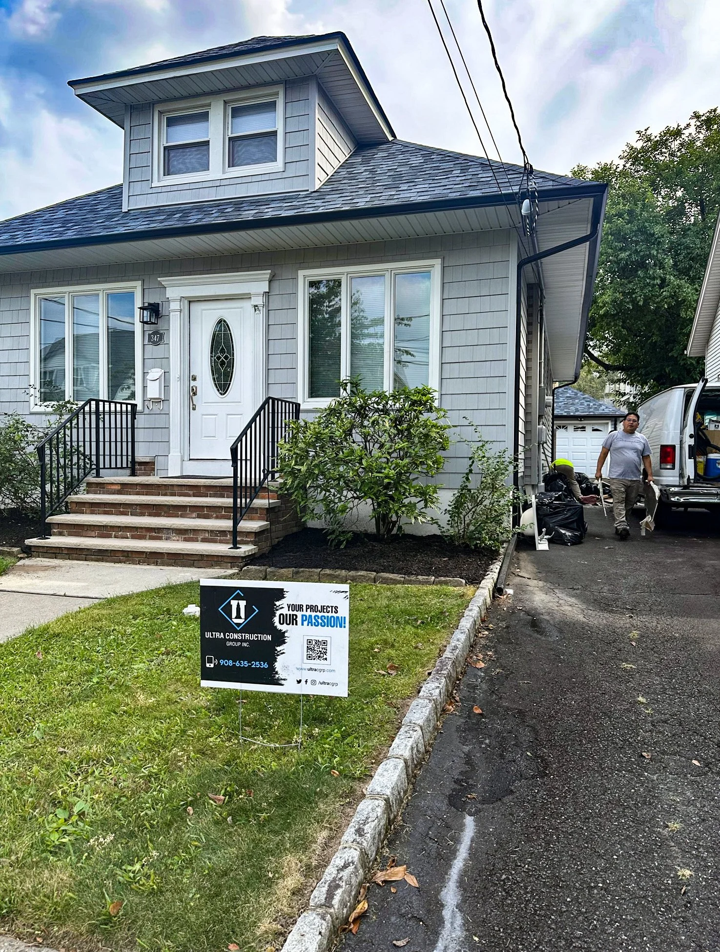A two-story house with light gray siding, a white front door with an oval glass window, brick steps, and black railings. There is a sign in the front yard advertising Ultra Construction Group, and two workers near a white van in the driveway.