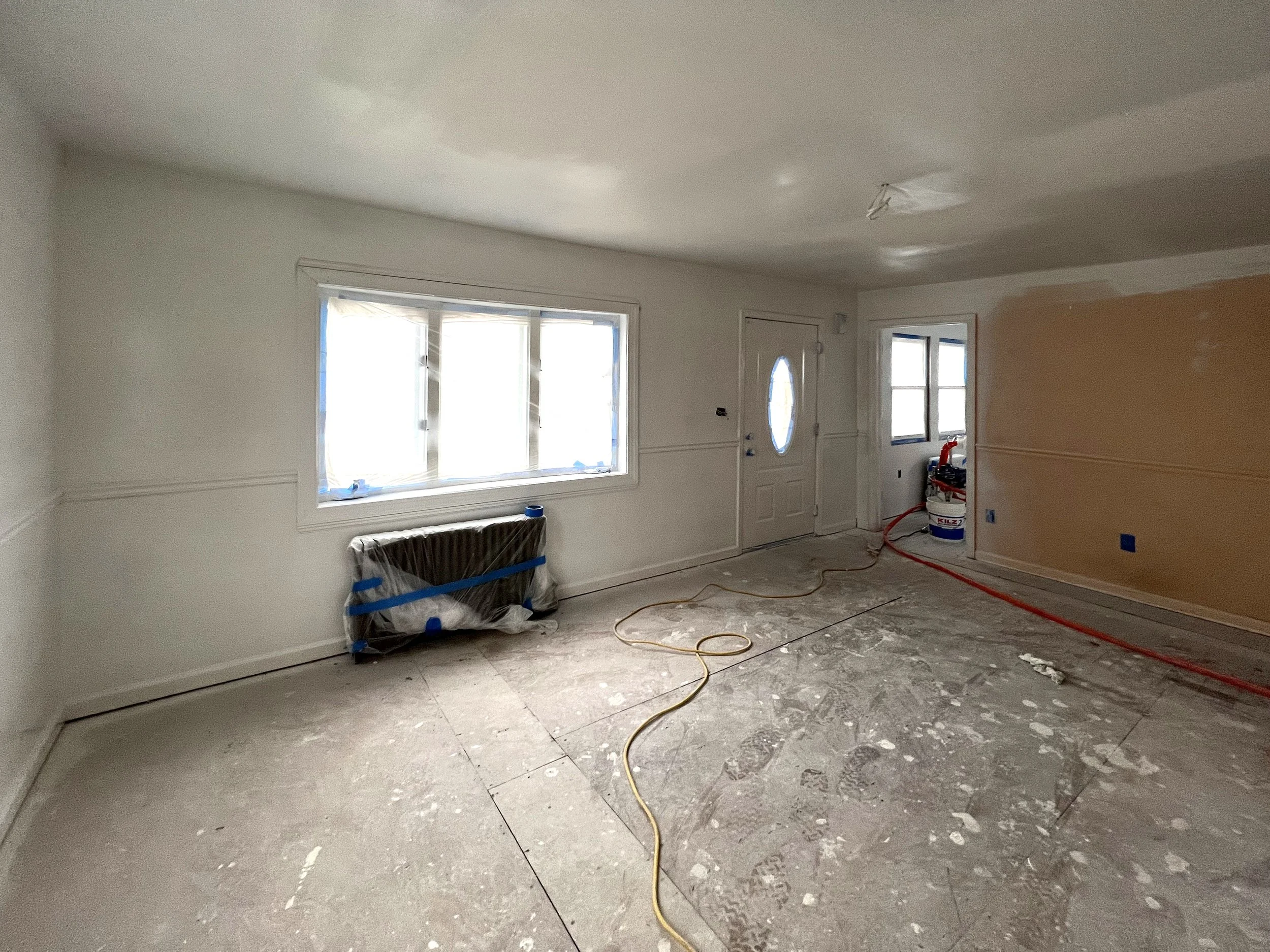 Living room under renovation with unpainted walls, covered window, construction tools, and exposed floor.