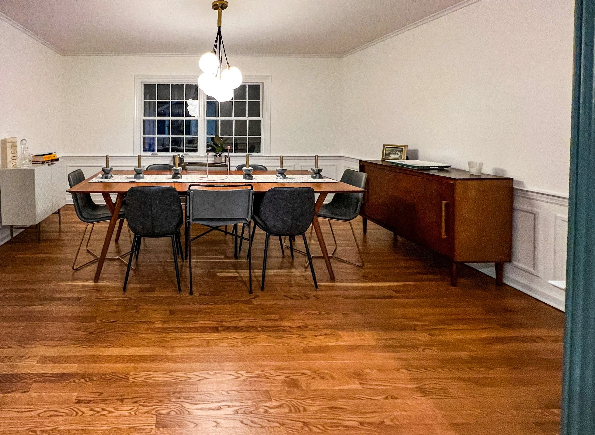 A dining room with a wooden table, eight black and gray chairs, a modern light fixture hanging above, a window on the back wall, a wooden sideboard on the right, and a white paneled wall with crown molding.