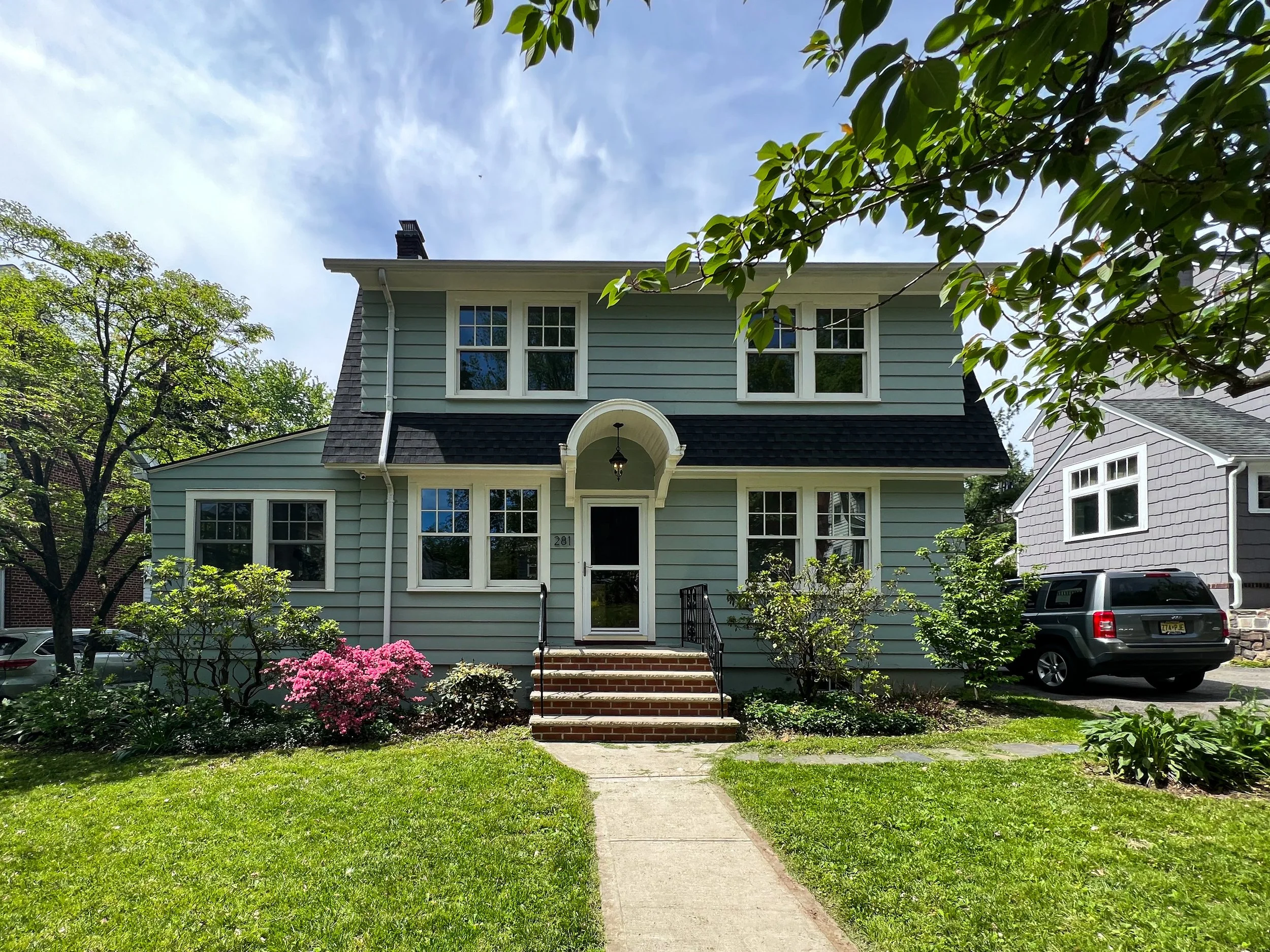 Front view of a light blue two-story house with white trim, a black roof, and a small front porch with brick steps. Green lawn and bushes surround the house, with trees and a sky with scattered clouds in the background.