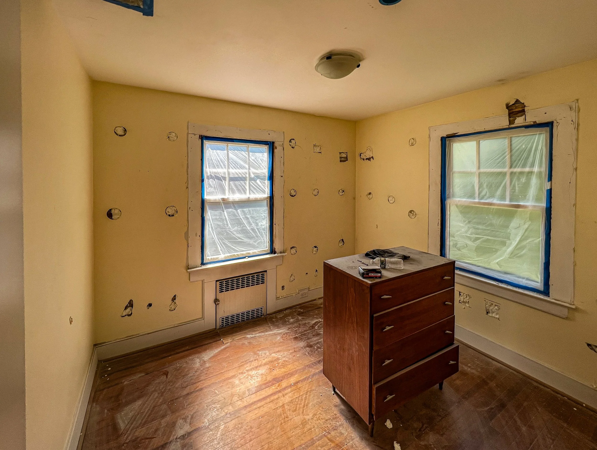 A room under renovation with yellow walls, two windows covered in plastic and tape, a wooden dresser in the center, and holes in the walls for future electrical work.