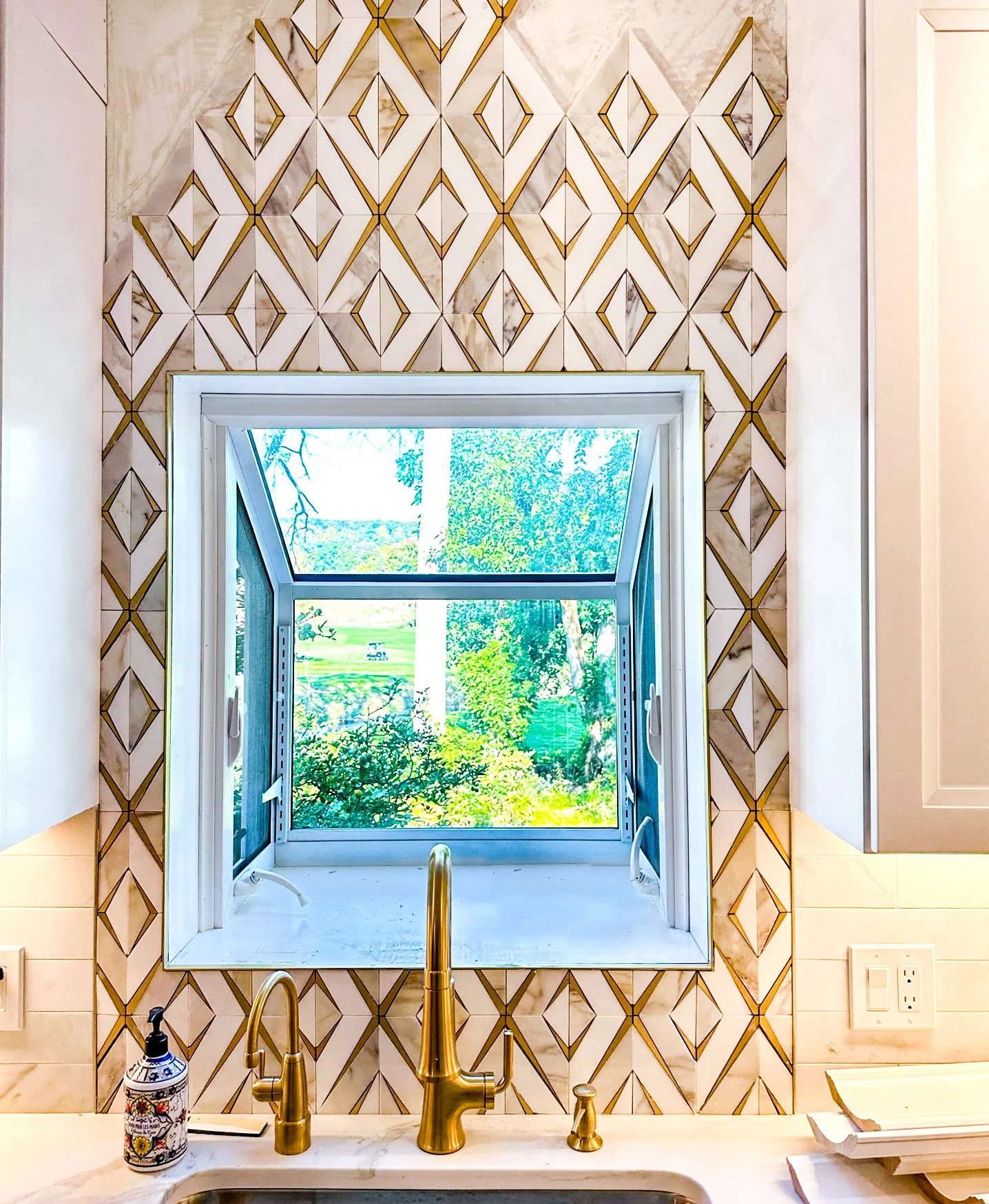 Kitchen window above the sink with a view of lush green trees and a golf course outside. The window has a tilt-open section at the top. The counter below has a golden faucet, a soap dispenser, and some kitchen items.