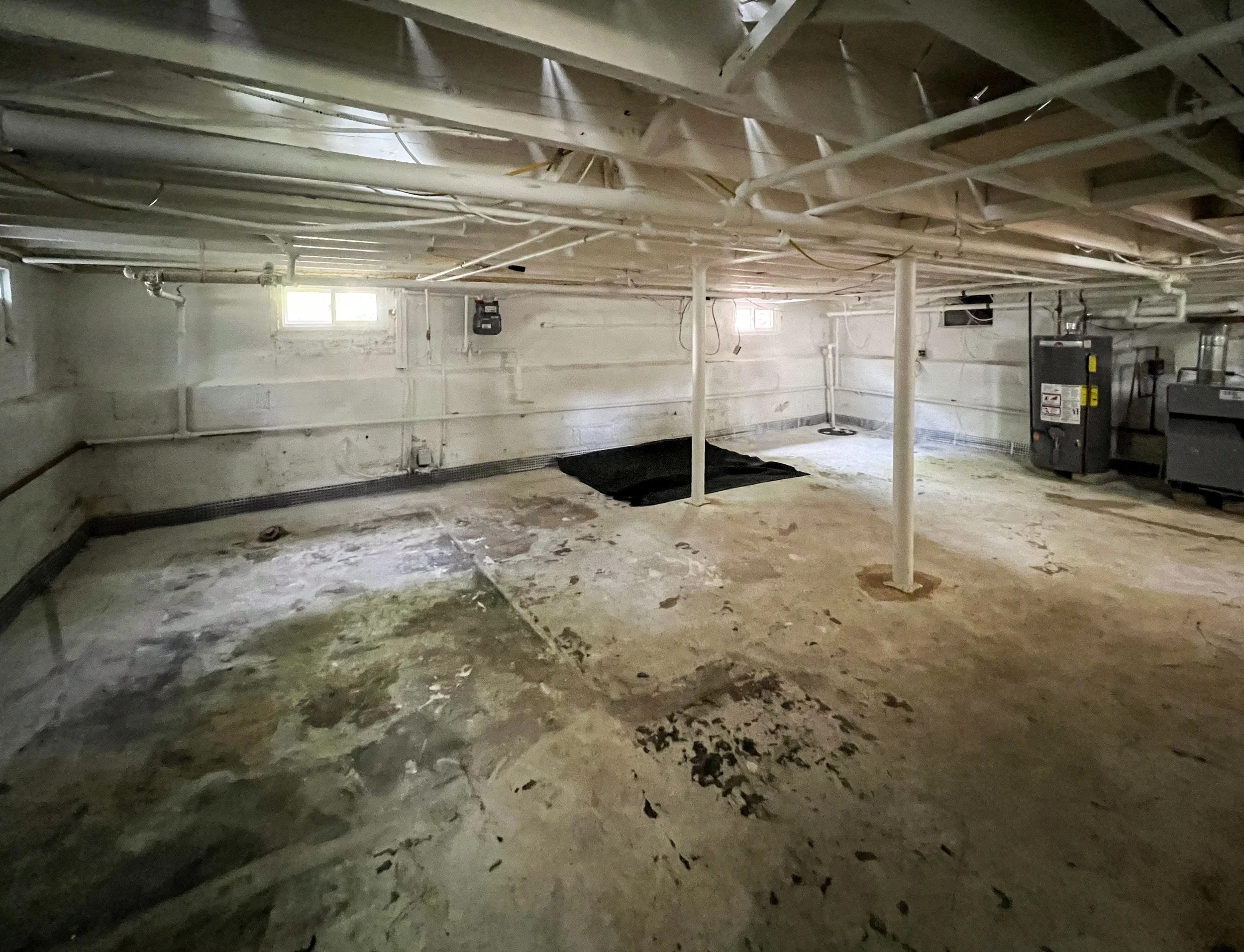 Empty basement with concrete floors and walls, pipes on the ceiling, small windows, and mechanical equipment in the corner.