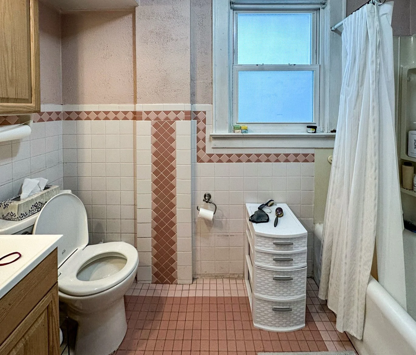 A small bathroom with pink and white tiles, a toilet, a window, a white storage drawer with grooming tools, a bathtub with a shower curtain, and bathroom supplies on a shelf.