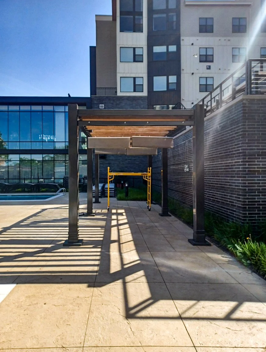 Outdoor area with a pergola, construction scaffold, and building in the background, showing shadows on the ground during daylight.