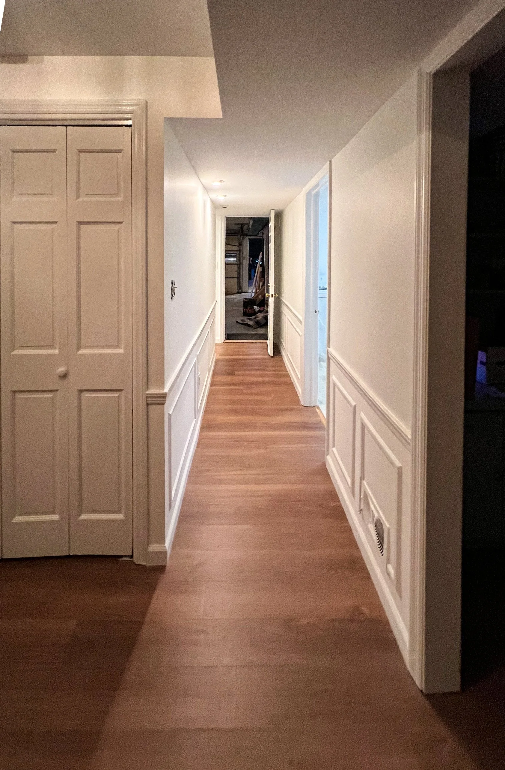 A hallway with wood flooring, white walls with decorative paneling, and open doorways leading to rooms. The hallway appears to be in a house under renovation or construction.