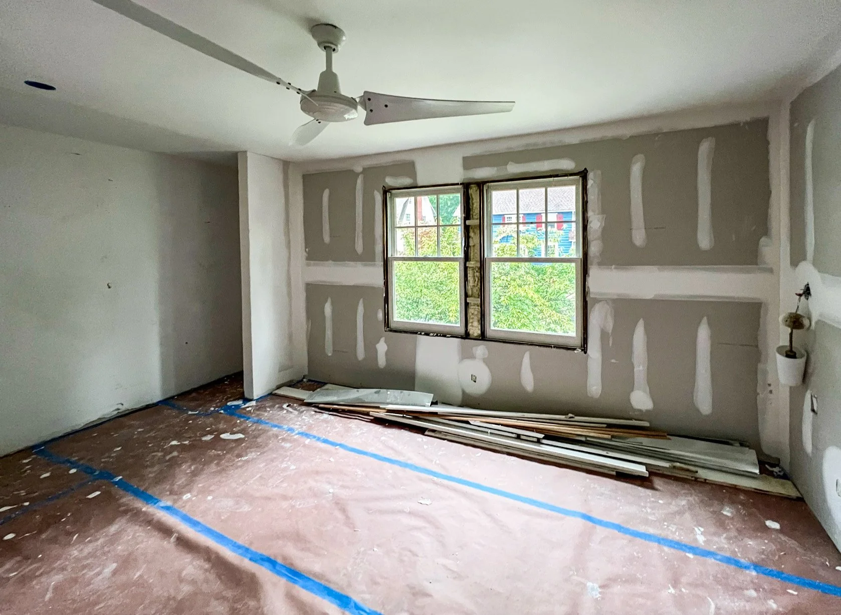 Empty room under construction with drywall panels and blue painter's tape on the floor, ceiling fan, and two windows showing a view of trees and neighboring buildings.