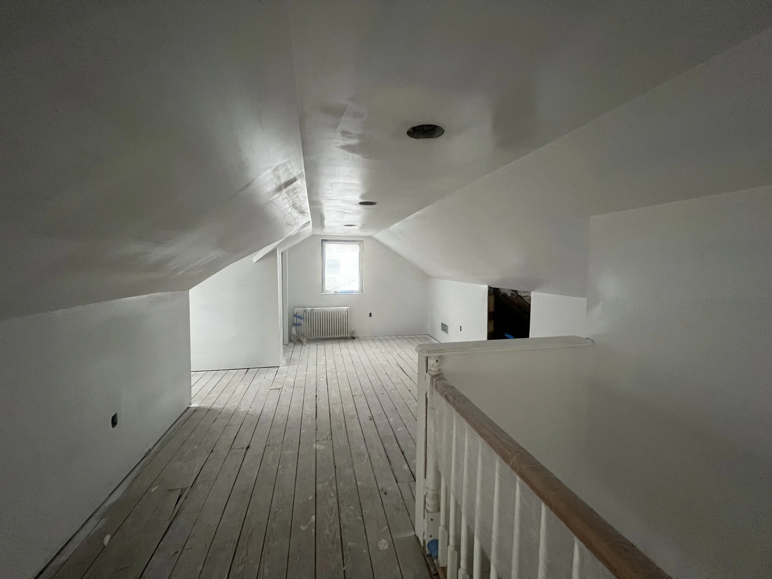 Empty attic room with sloped ceiling, white walls, a small window, and unfinished wooden flooring.