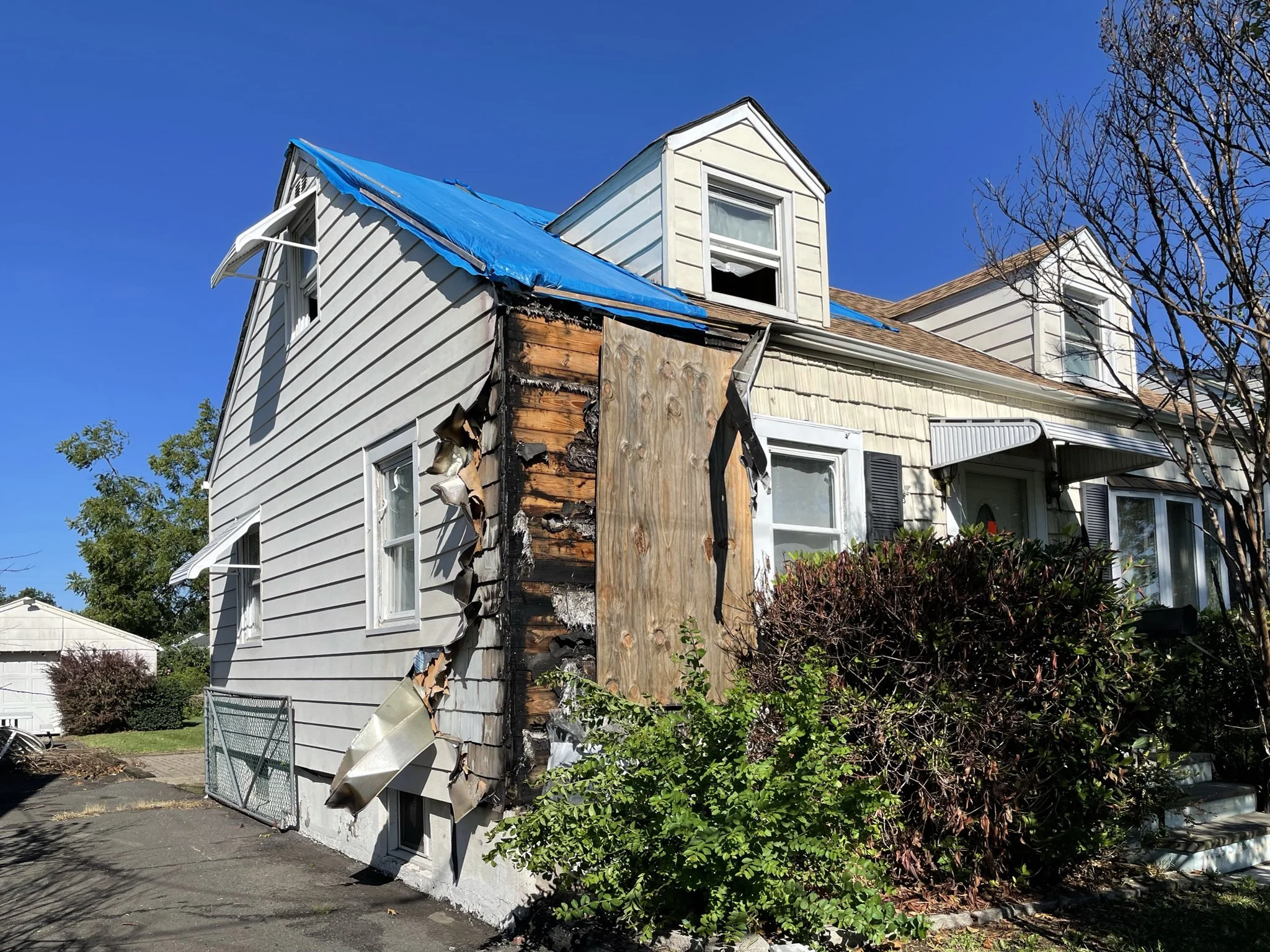 A damaged two-story house with burned exterior wall and peeling siding, blue tarp replacing part of the roof, neighboring house visible, and a bush in front.